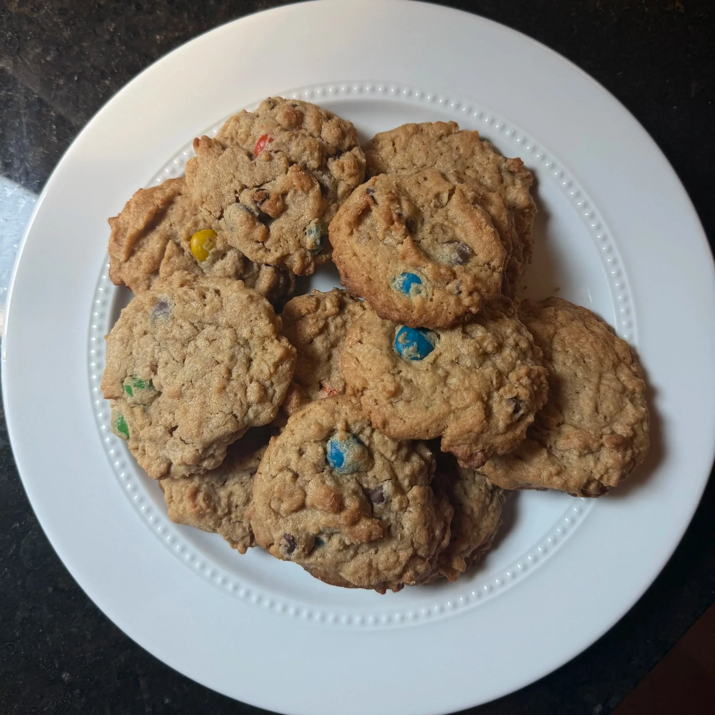 Plate of homemade monster cookies made with chocolate chips and colorful candy-coated chocolates. The golden brown cookies are stacked on a white plate, showcasing their soft, chewy texture