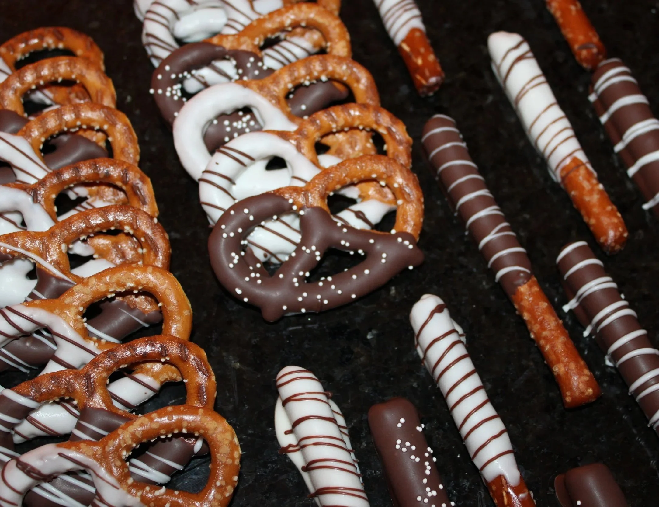 Assorted chocolate-covered pretzels and pretzel rods decorated with milk and white chocolate drizzles. The sweet treats are arranged on a dark surface and finished with white sprinkles for an elegant homemade dessert display.