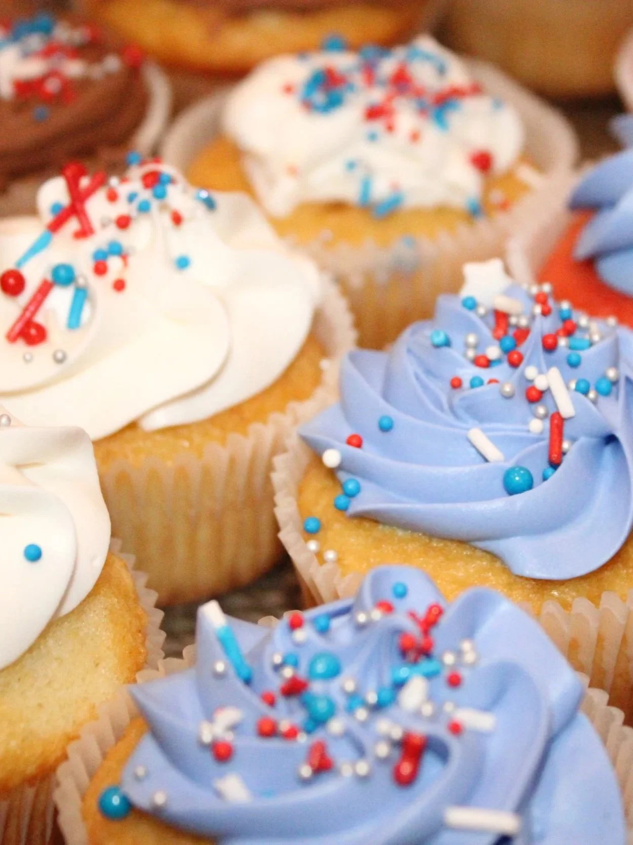 Assorted vanilla and chocolate cupcakes topped with white and light blue buttercream swirls and finished with red, white, and blue sprinkles. The cupcakes are arranged together in a festive patriotic display.