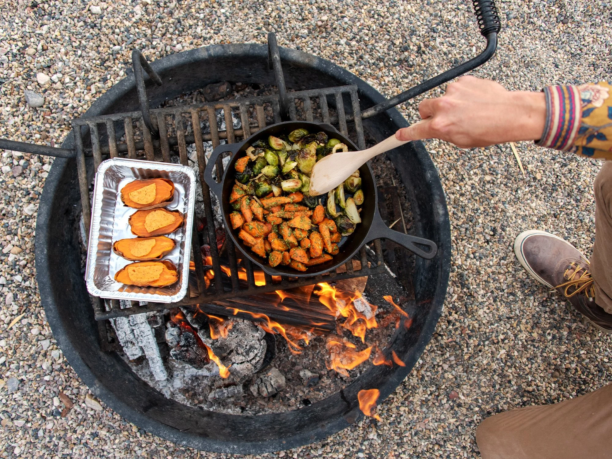 man stirring grilled vegetables in a cast iron pan over a wood burning fire