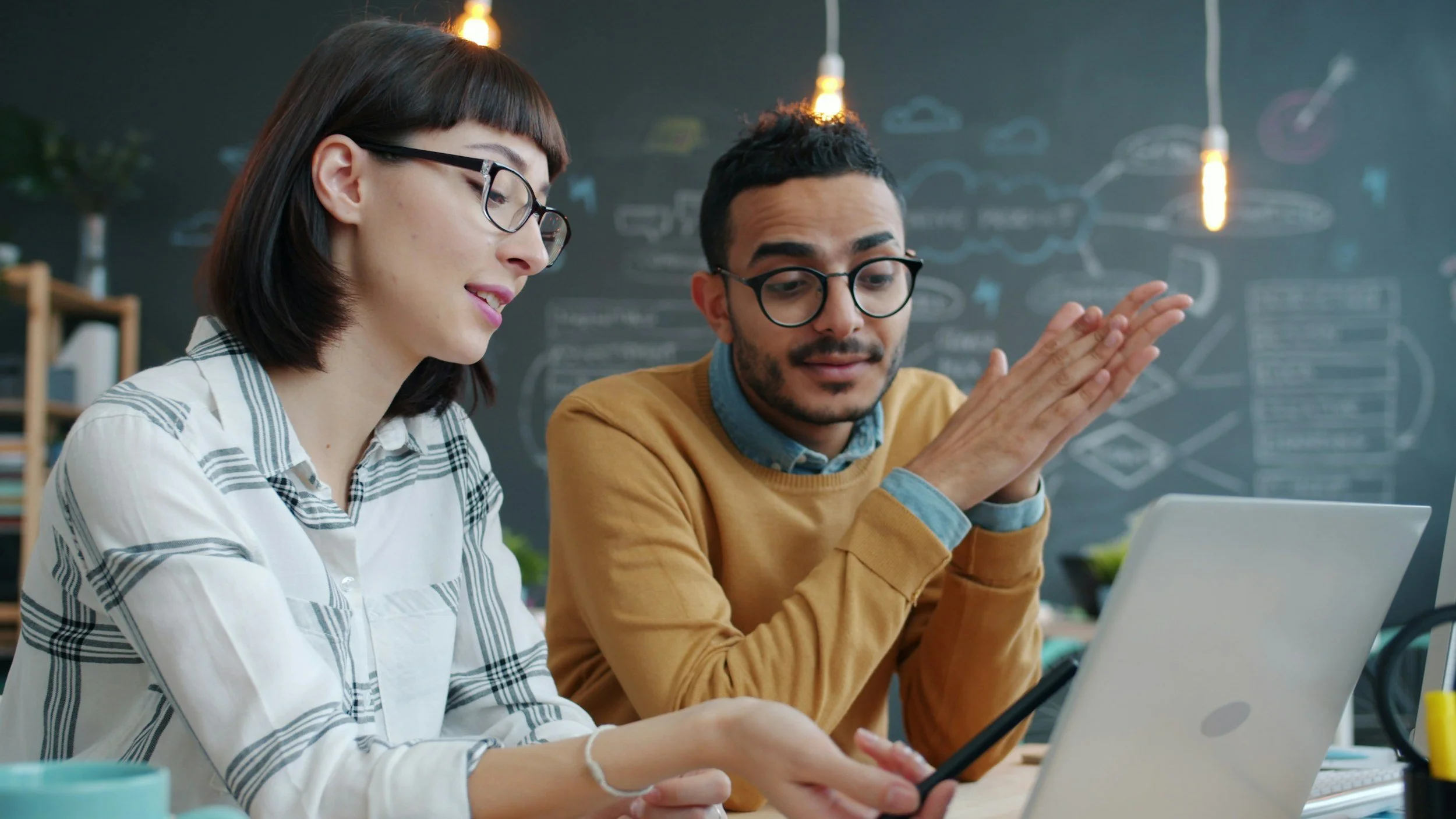 Two diverse young adults, a woman with glasses and a man with dark hair and glasses, are collaborating on a project using a tablet in a modern office with a chalkboard wall.
