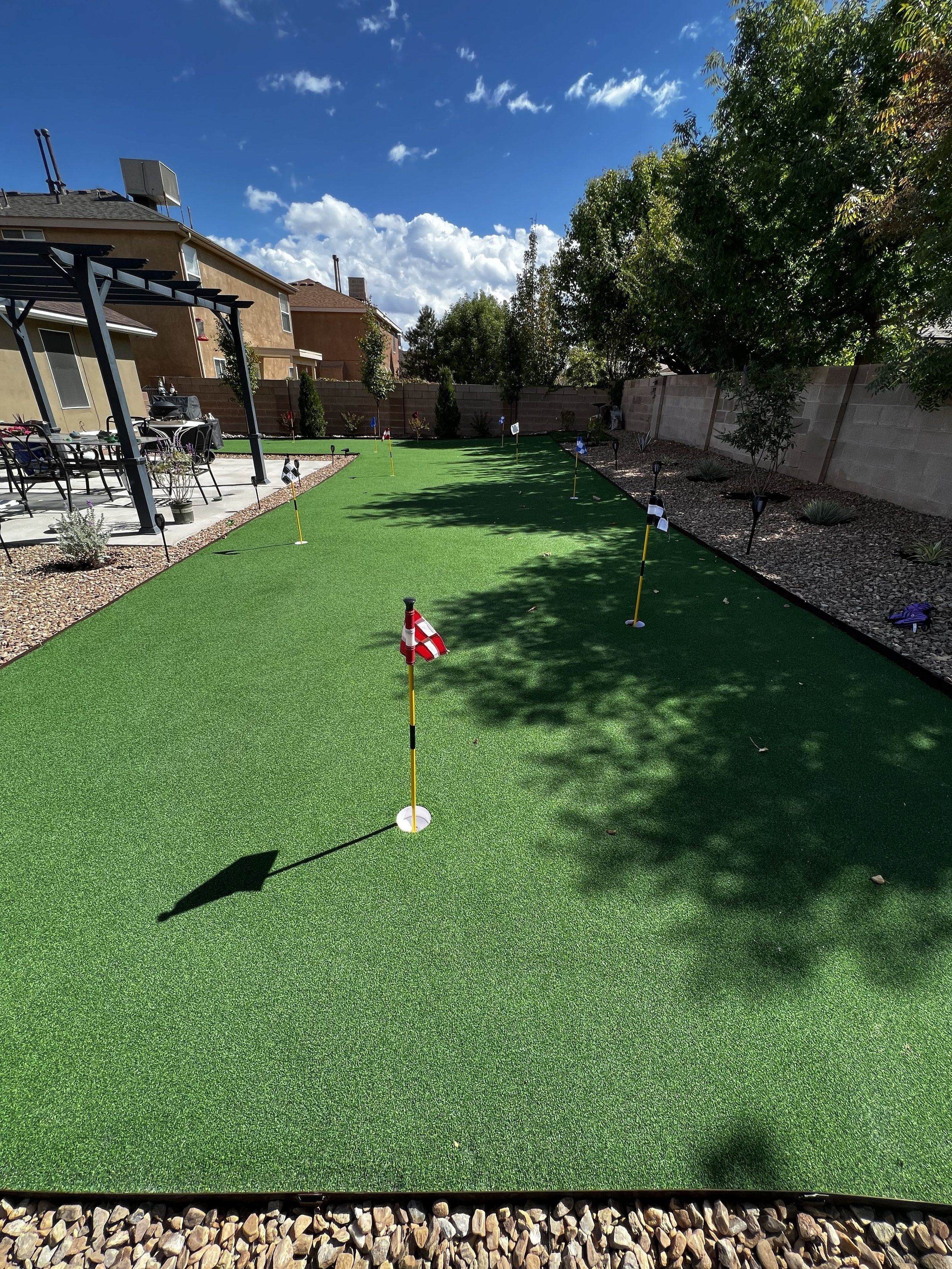 A backyard with artificial grass and mini golf flags, a patio with outdoor furniture, and a tall tree casting shadows, under a partly cloudy blue sky.
