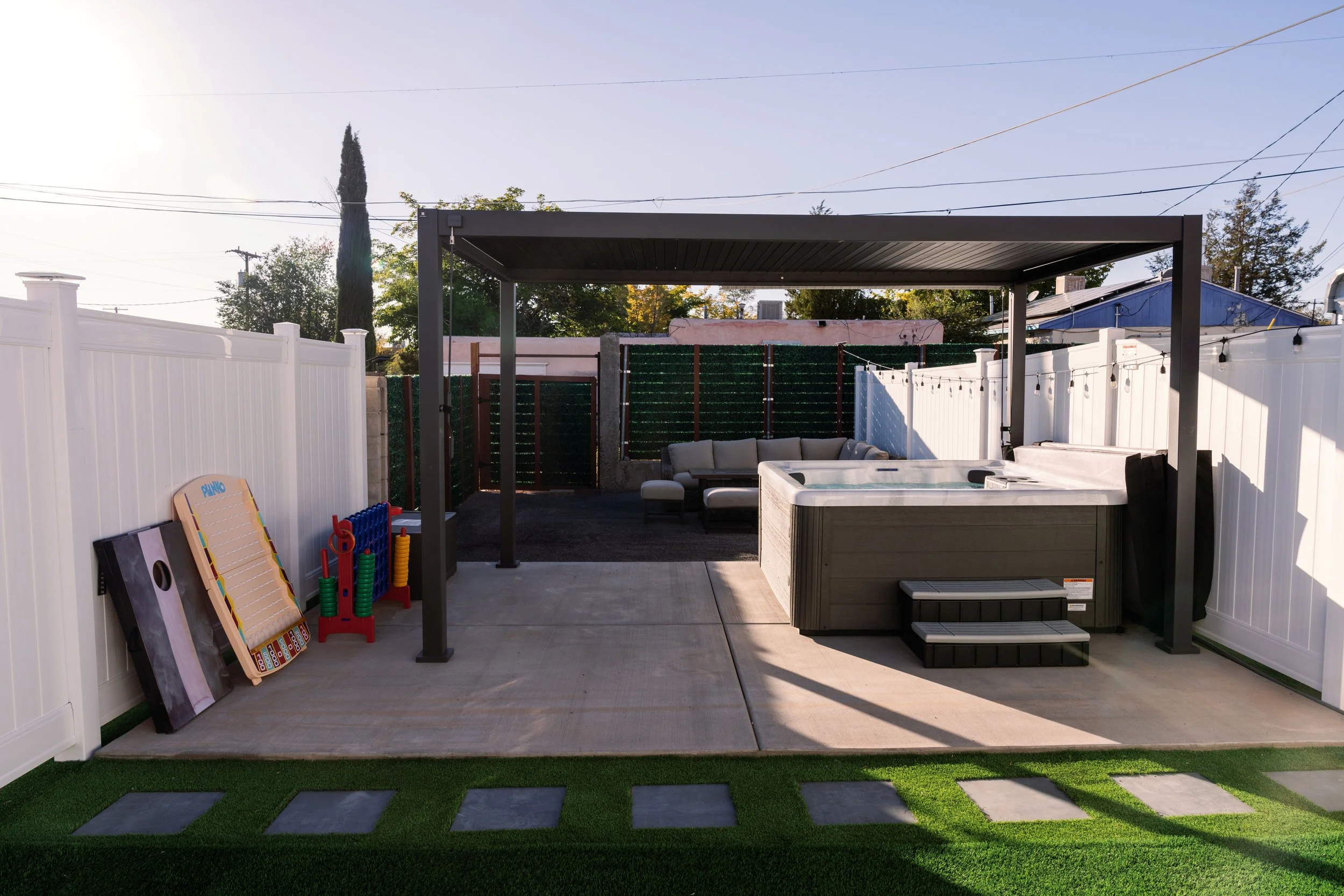 A backyard patio with a covered seating area, white fencing, children's toys, a hot tub, and a patch of artificial grass.