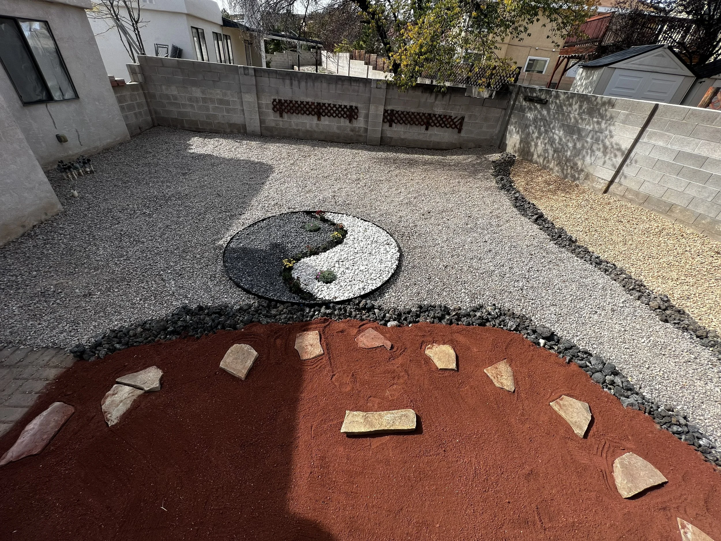 A backyard with a concrete block wall, a small garden bed with white gravel and plants in a yin-yang shape, flanked by red mulch and stone borders, and a gravel ground surface.