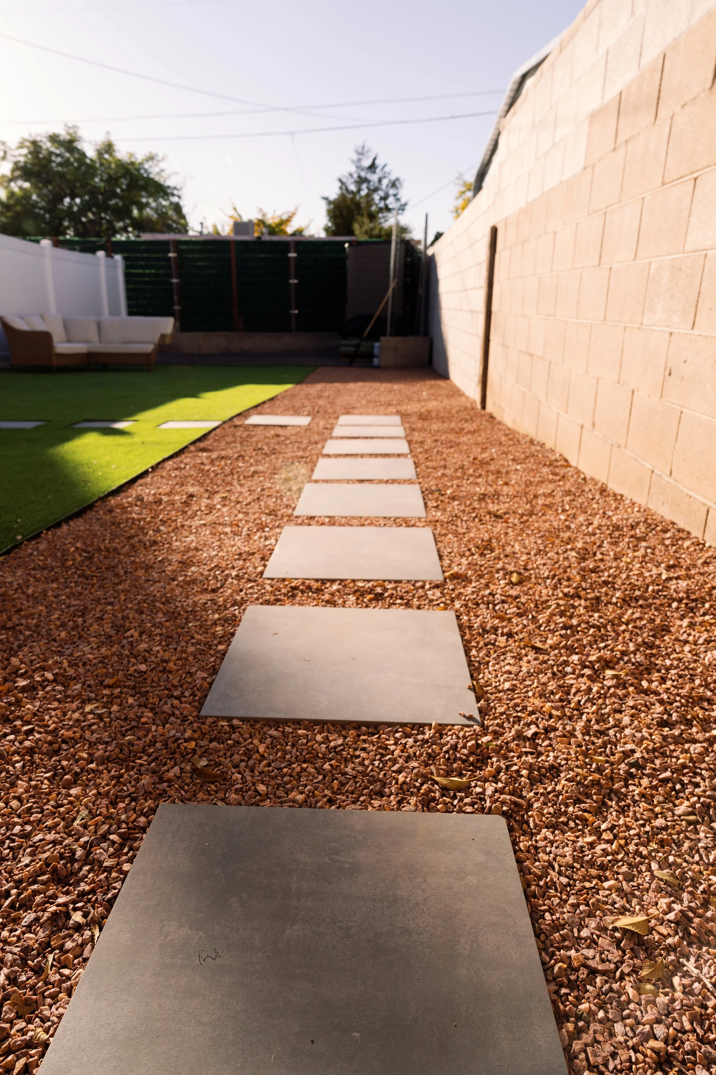 A pathway consisting of large square concrete stepping stones runs through a garden with reddish-brown gravel on the sides, leading towards a wall made of large beige bricks on the right, and a patch of green artificial grass on the left.