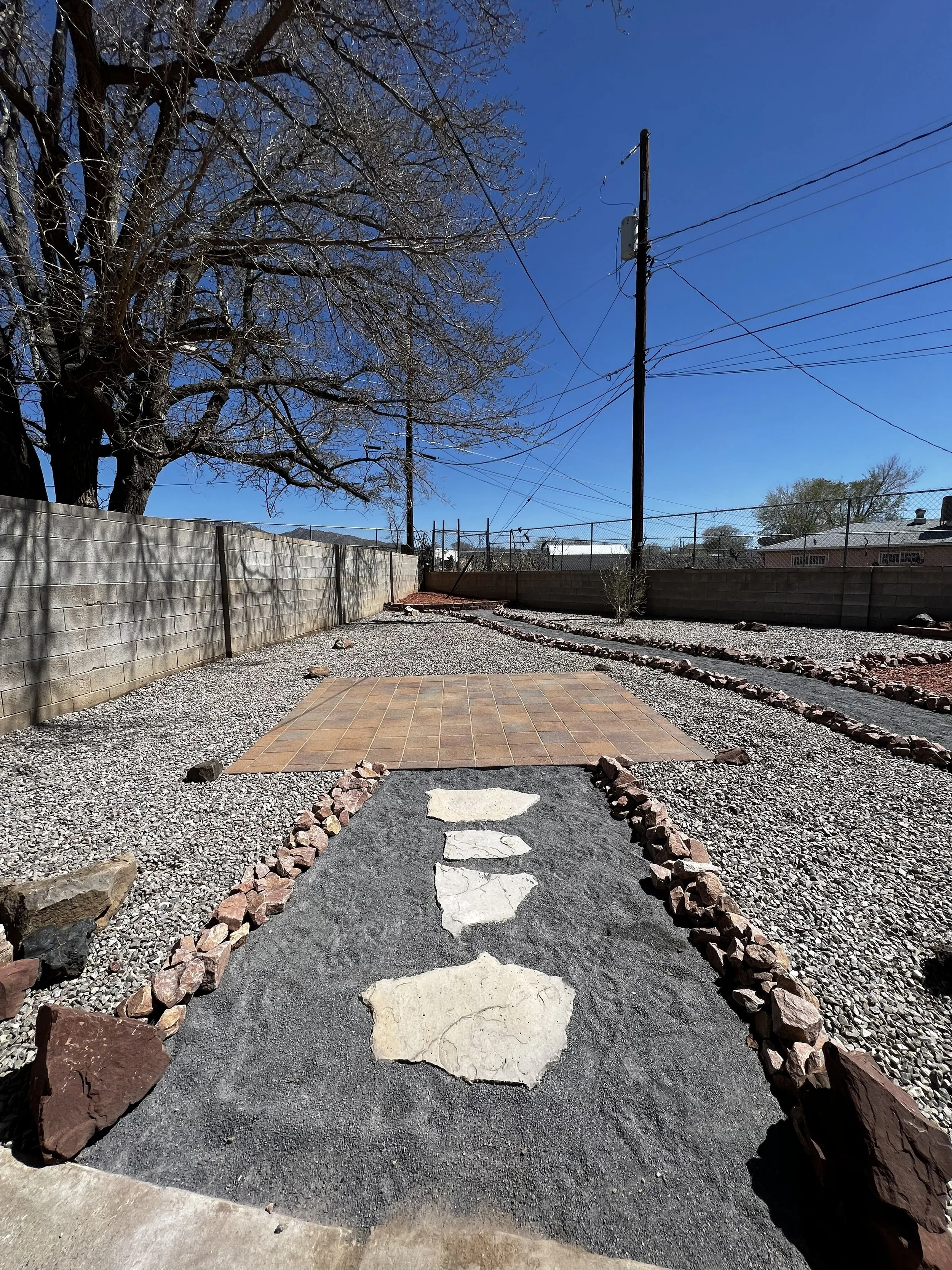 A backyard with a gravel path bordered by rocks leading to a tiled area, enclosed by a concrete block wall, with a large tree and utility pole with power lines against a blue sky.
