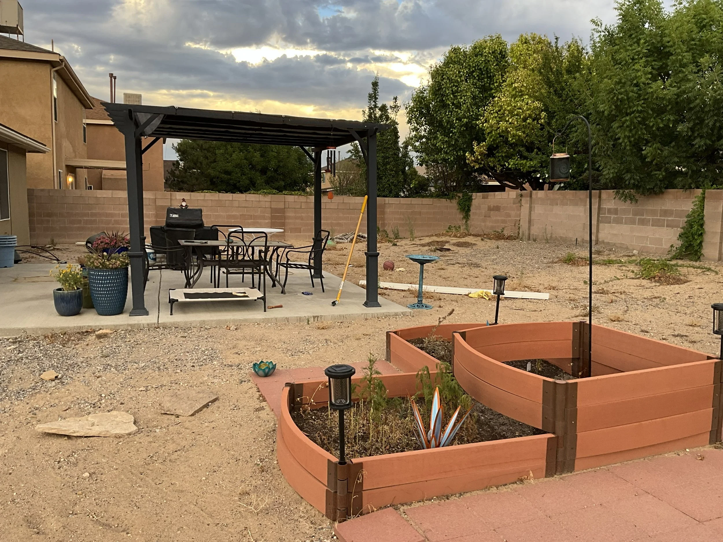 A backyard with a covered patio area featuring a table and chairs, potted plants, a garden bed with a small plant, and a new planting area with brick borders. There is a brick wall, trees, and a cloudy sky in the background.