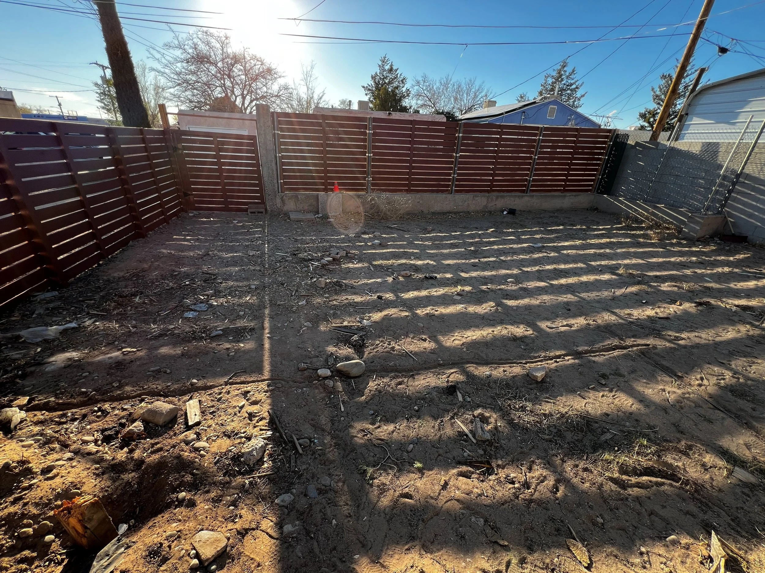 Empty backyard with dirt ground, scattered rocks, a newly installed brown wooden fence, and sunlight shining through the trees.