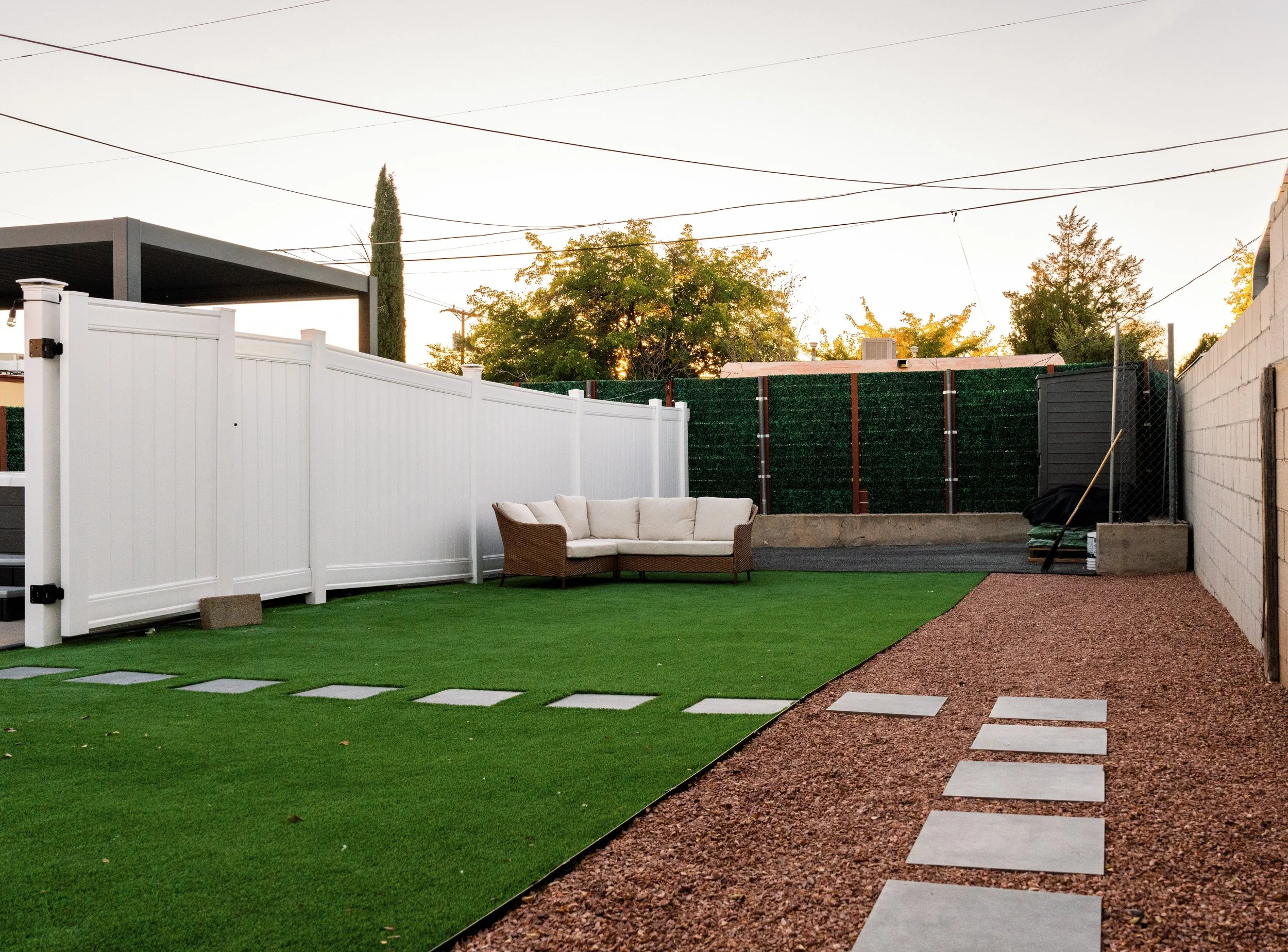 Backyard with white privacy fence, artificial grass, and concrete stepping stones leading to a patio with outdoor seating. Trees and a fence are visible in the background, with power lines overhead during sunset.