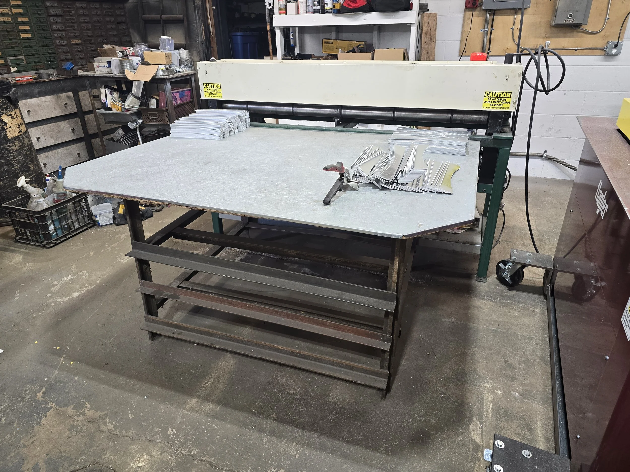 A workshop with a large metal work table in the center with stacks of thin metal sheets and a clamp on top. In the background, there are shelves with tools and supplies, and a machine to the right with warning labels. The floor is concrete with various equipment and materials stored around.