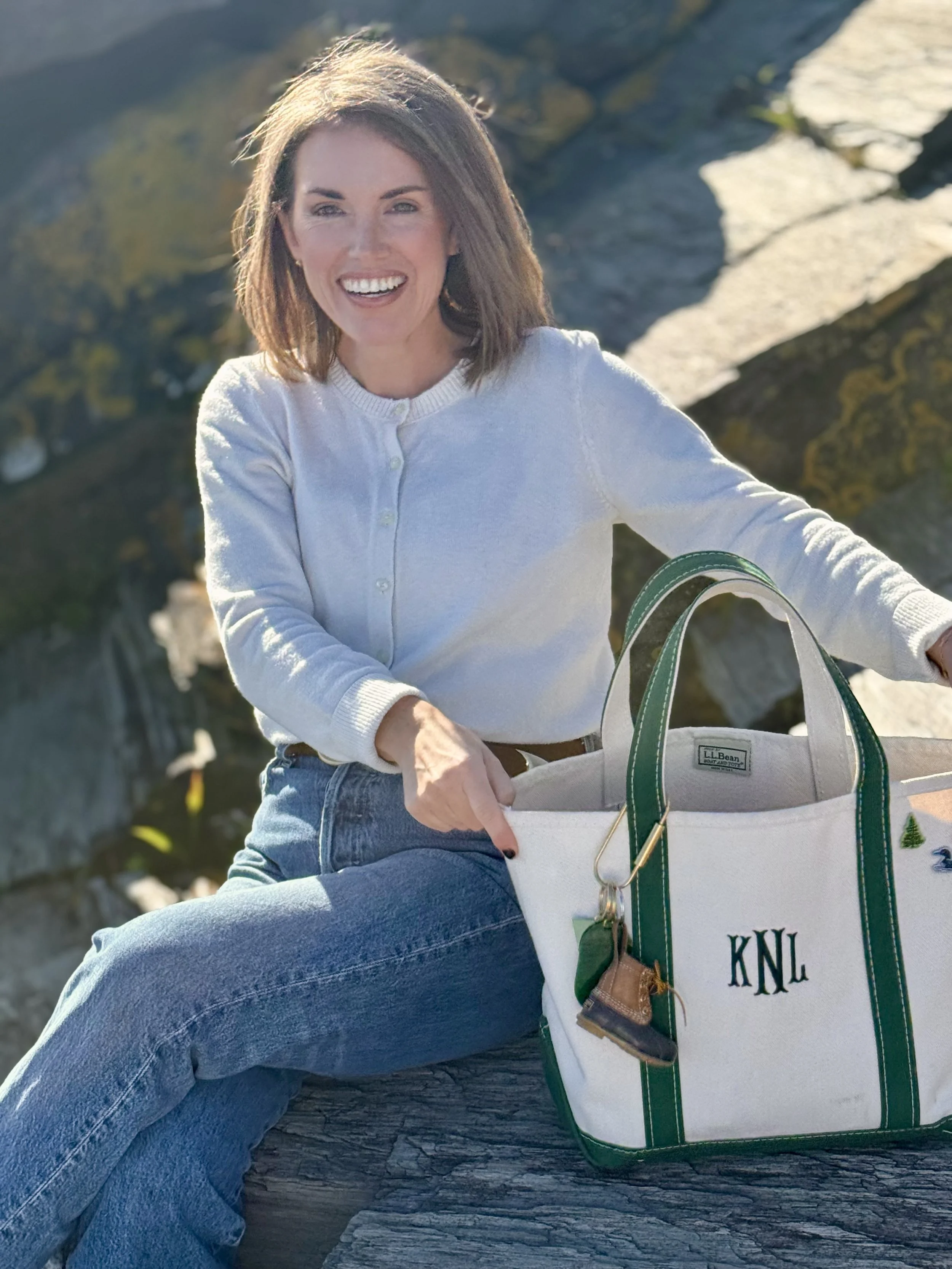Smiling woman in a light sweater sitting beside a lake with a monogrammed LL Bean tote bag.