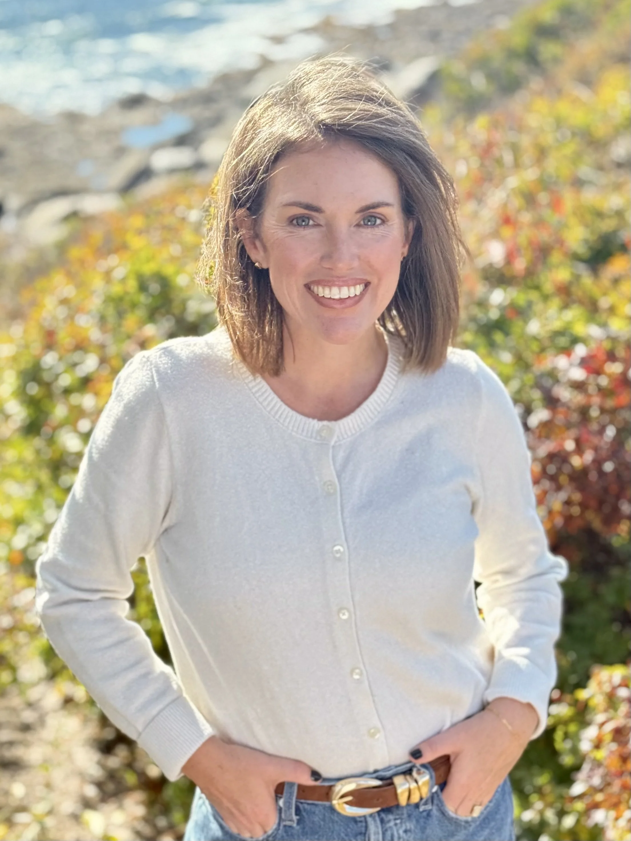 A woman with shoulder-length brown hair, wearing a white sweater, blue jeans, and a brown belt, standing outdoors with autumn foliage in the background.