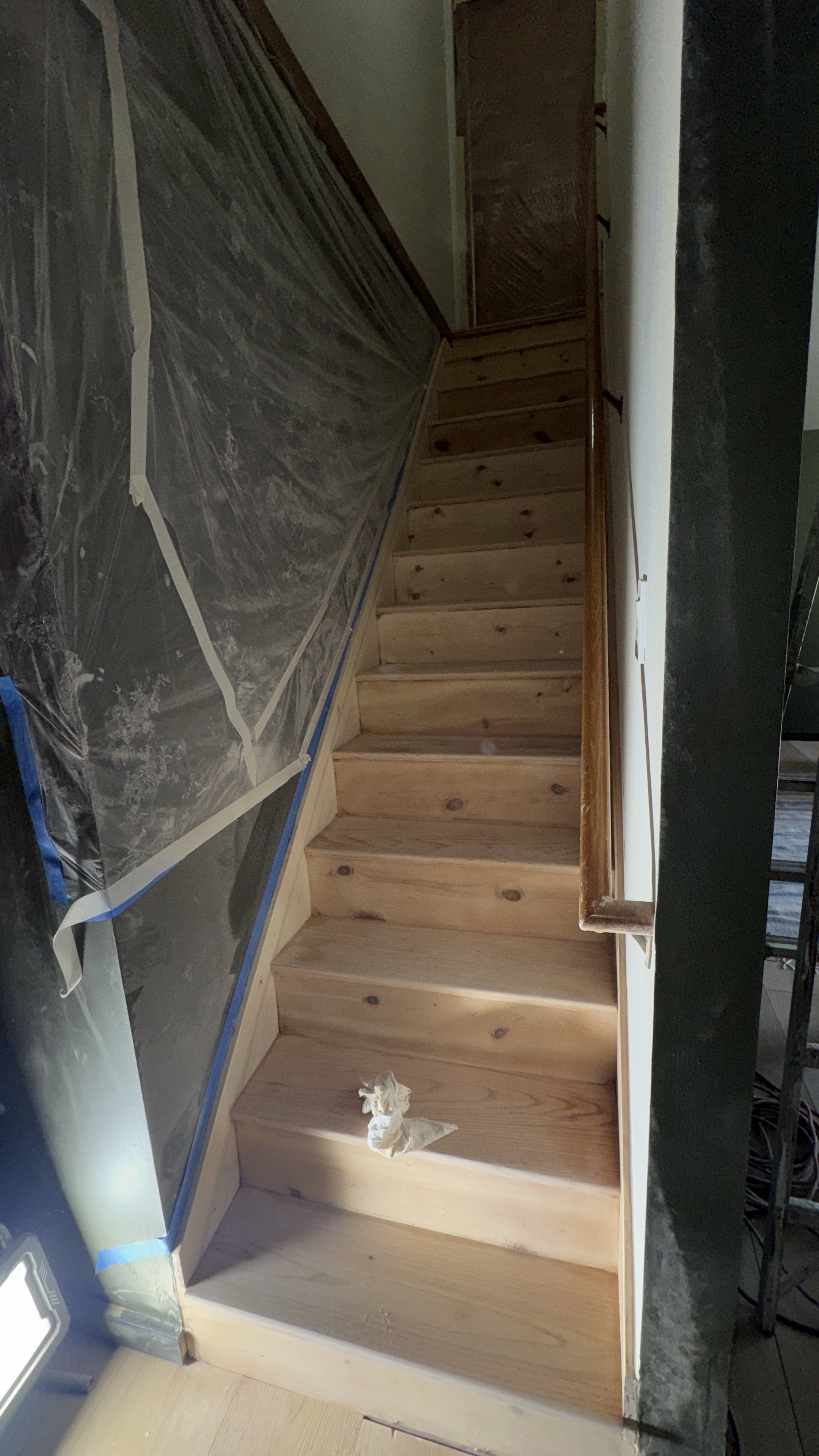 Wooden staircase under construction, with plastic sheeting on the wall to the left, and a black object on the right side of the image. There is a crumpled piece of tissue or cloth on the floor at the bottom of the stairs.