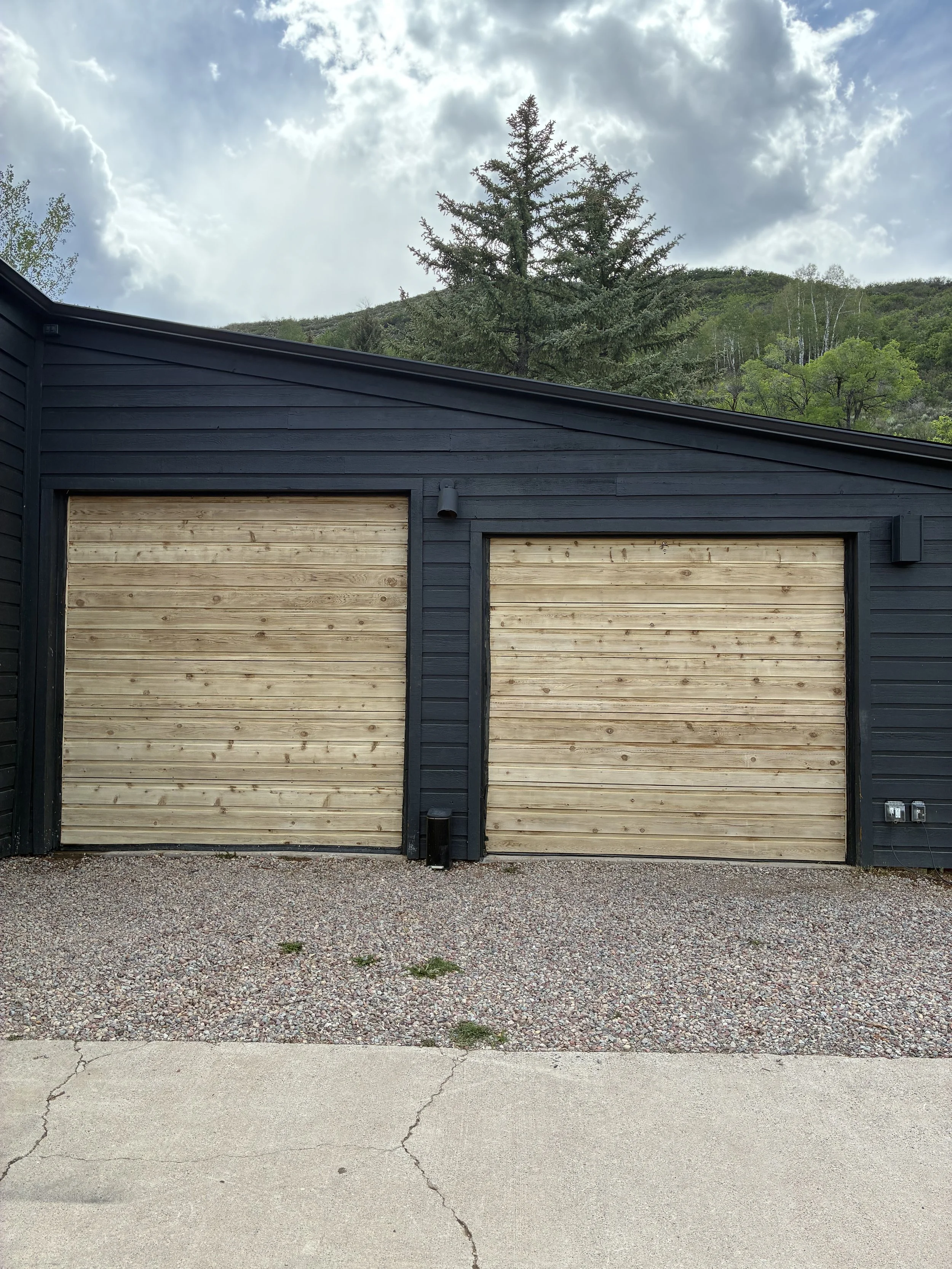 Photo of a black garage door with wooden panels, set against a dark-colored building with a sloped roof. The ground in front is a gravel driveway with some cracks in the concrete border at the bottom, and a backdrop of green trees and a cloudy sky.