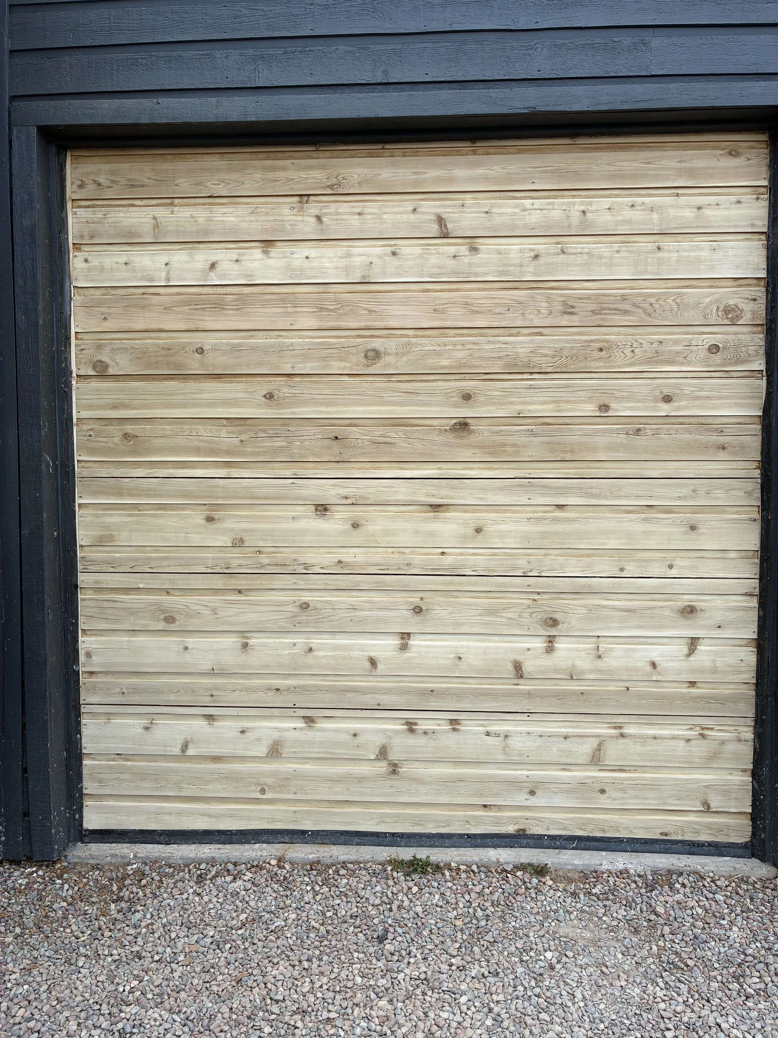 A closed beige wooden garage door with horizontal planks, framed by black trim, with a gravel driveway in front.