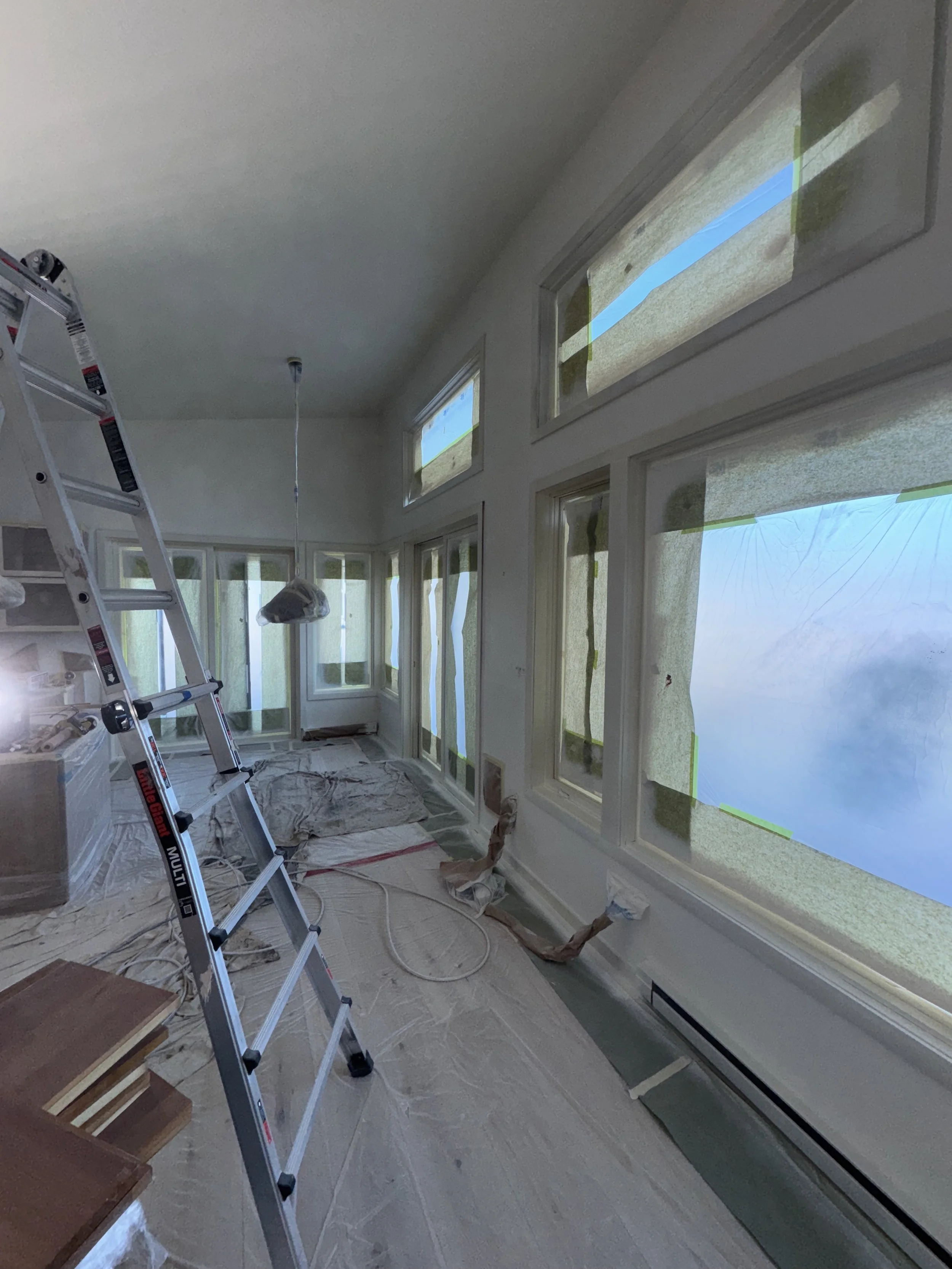 Interior room under renovation with large windows covered in protective tape, a ladder, and construction materials on the floor.