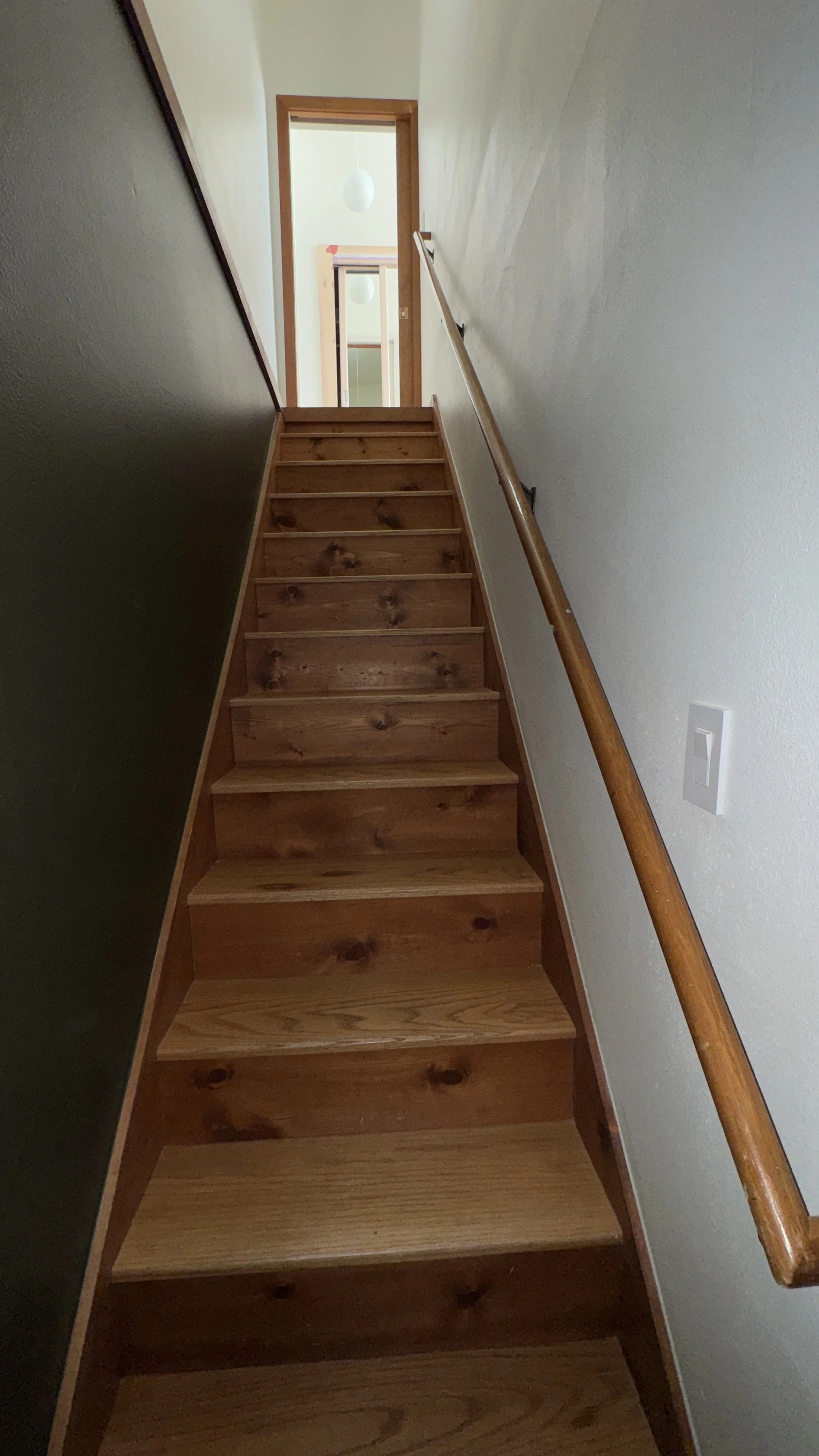 Wooden staircase with a handrail on the right side, leading up to a doorway with a wooden frame. Light fixtures are visible in the room beyond the doorway.