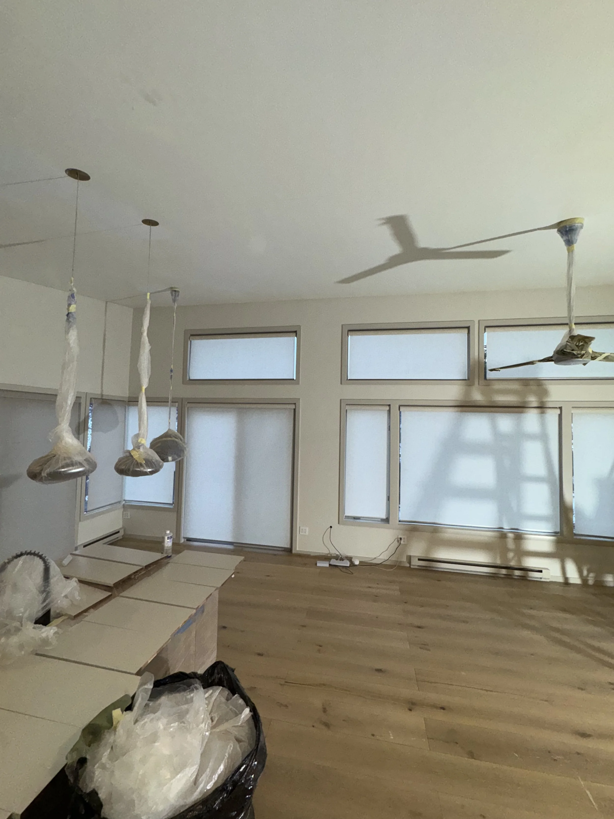A room under renovation with hanging light fixtures covered in plastic, large windows with frosted glass, and a wooden floor. Construction supplies and tools are scattered around.