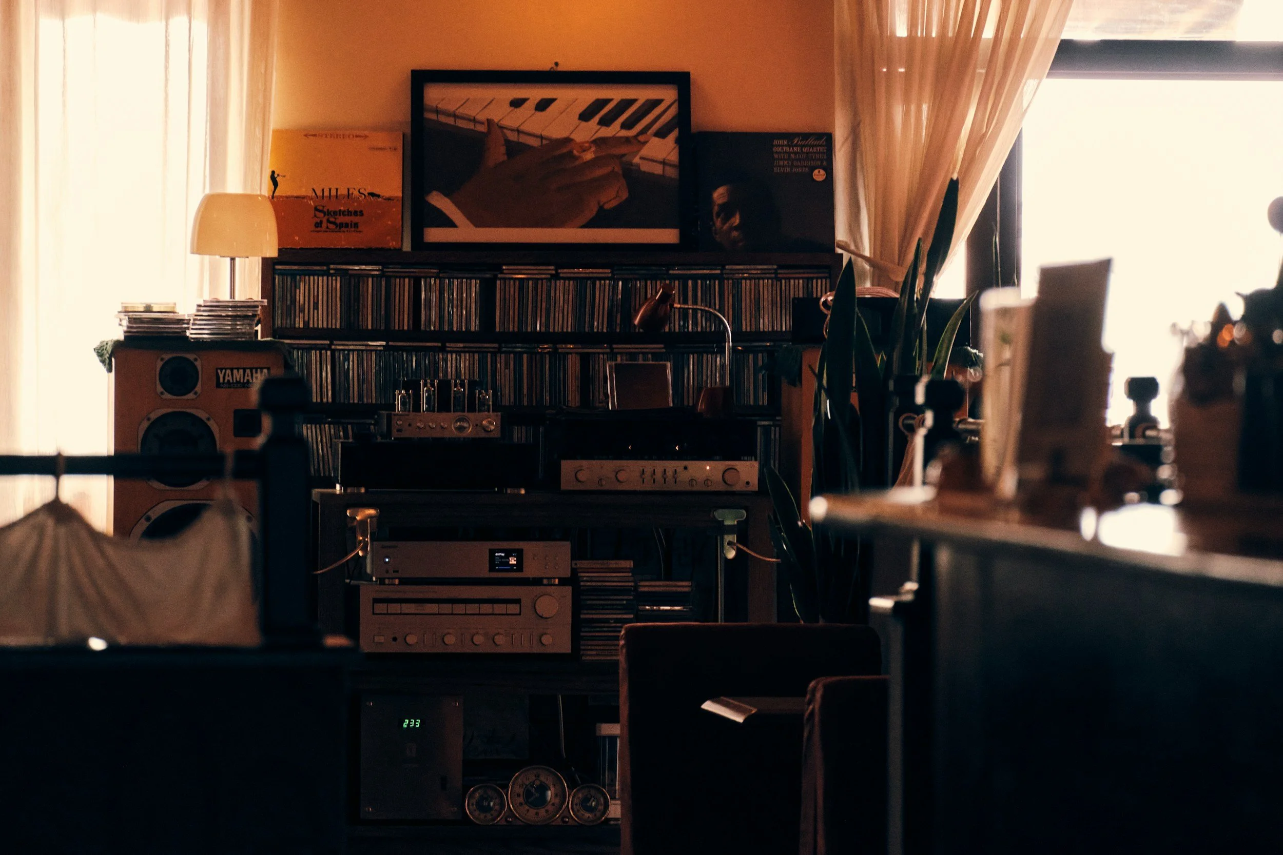 Interior of a room with a shelf holding vinyl records and music equipment, framed artwork on the wall, a lamp, and a large window with curtains allowing natural light to illuminate the space.