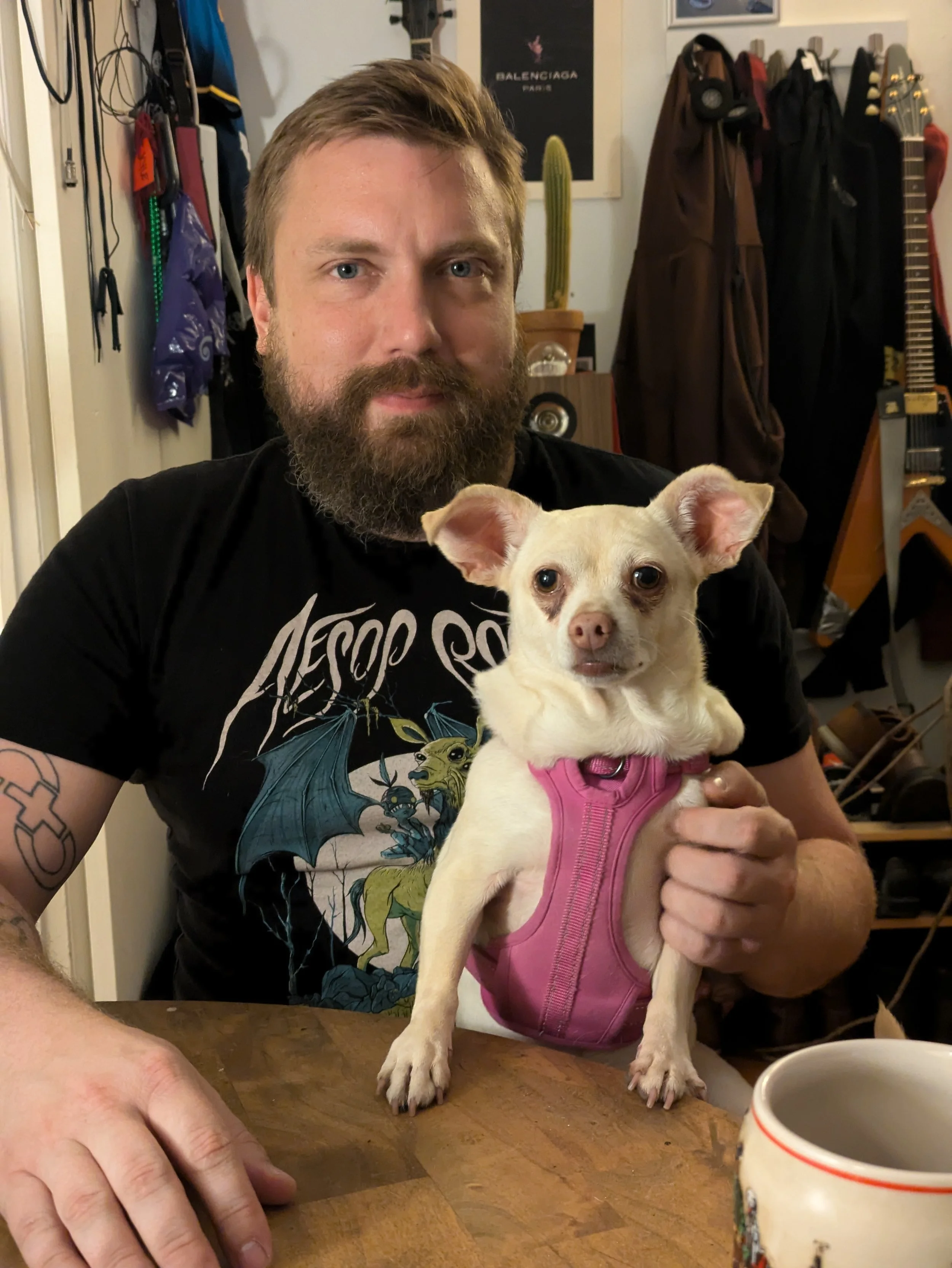 A man with a beard and a tattoo on his arm is holding a small tan Chihuahua dog wearing a pink harness. They are indoors at a wooden table with a cup in front of them. The background shows coats hanging, a cactus in a pot, and posters on the wall.