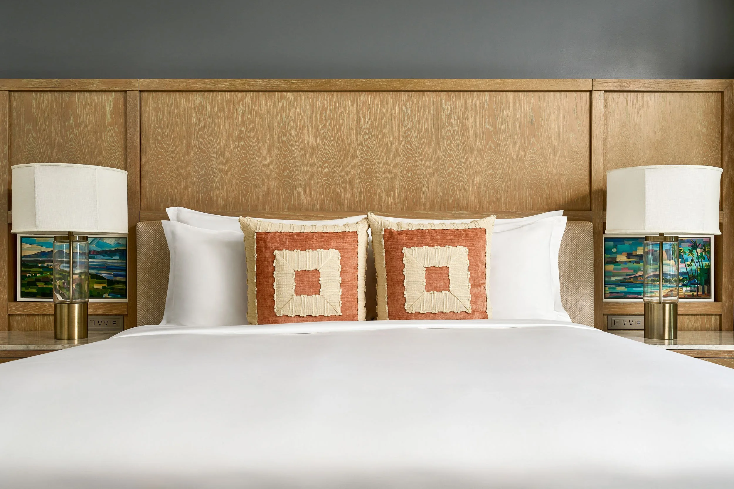 A neatly made bed with white sheets and two decorative pillows with a geometric pattern, flanked by matching lamps on side tables, against a wooden headboard and a gray wall.