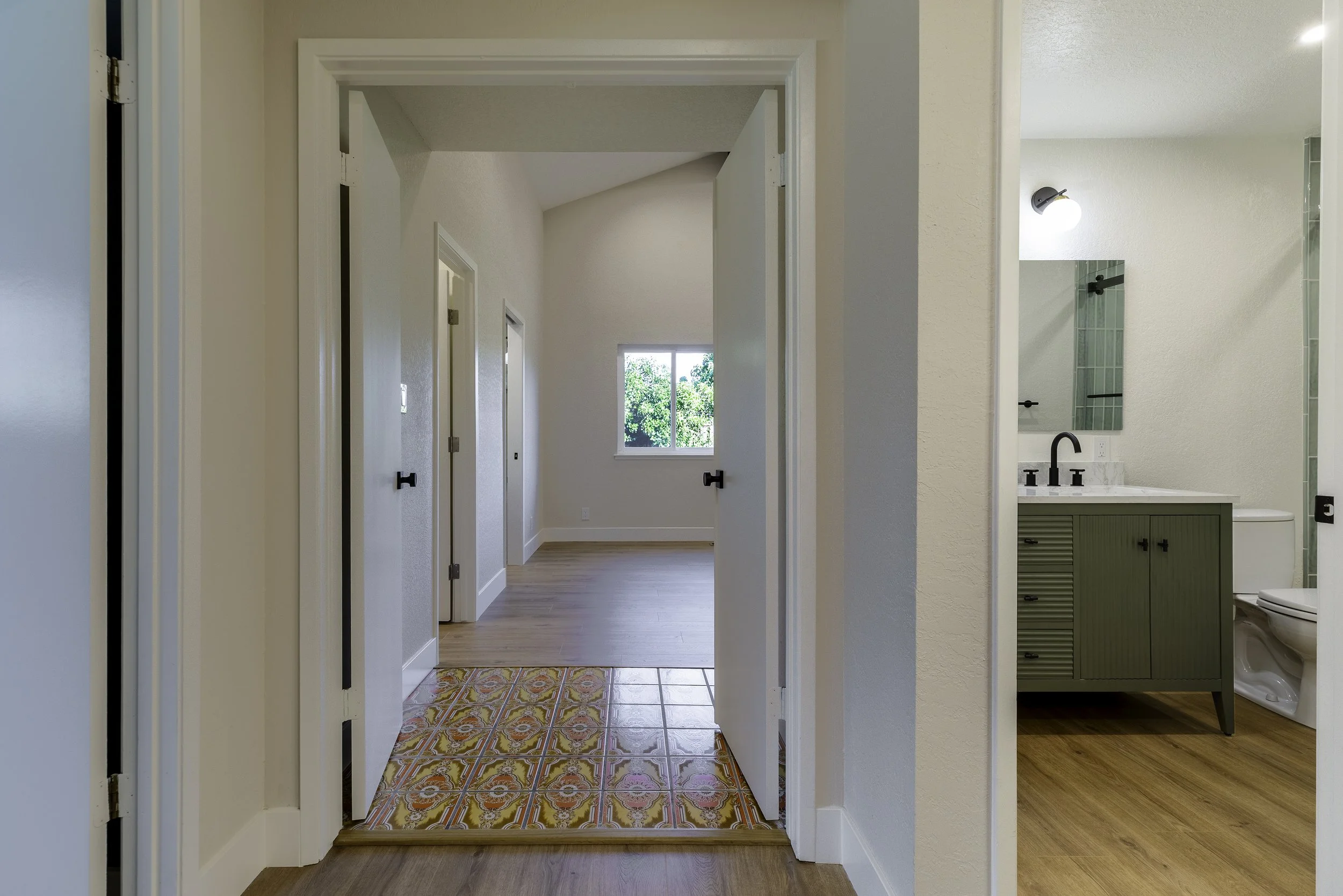 View of a hallway with patterned tile flooring leading to a brighter room with a large window and a smaller room with a bathroom on the right.