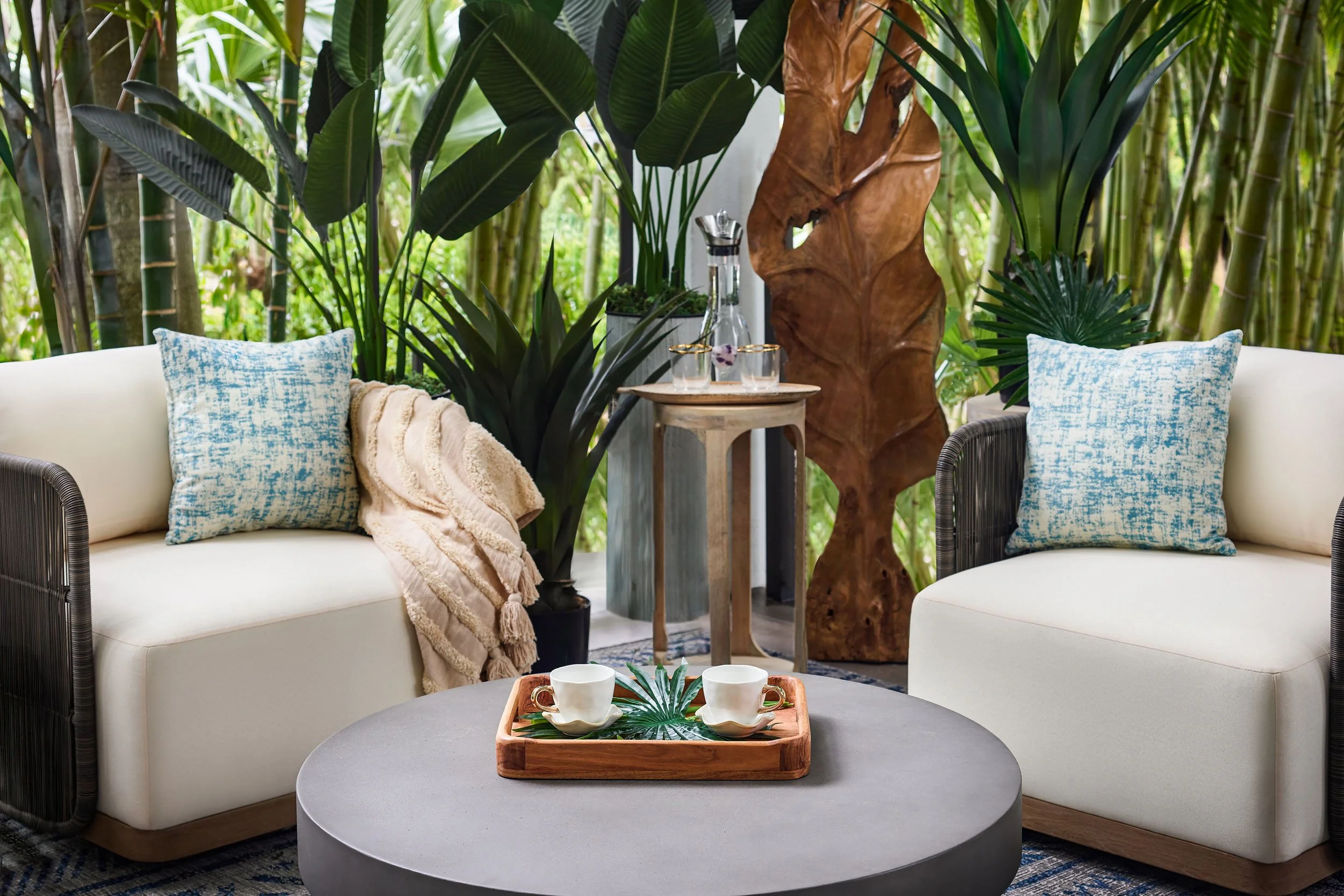 Living room area with two white sofas with blue and white patterned pillows, a gray round coffee table with a wooden tray, and green plants including bamboo, banana, and palm, with a wooden sculpture in the background.