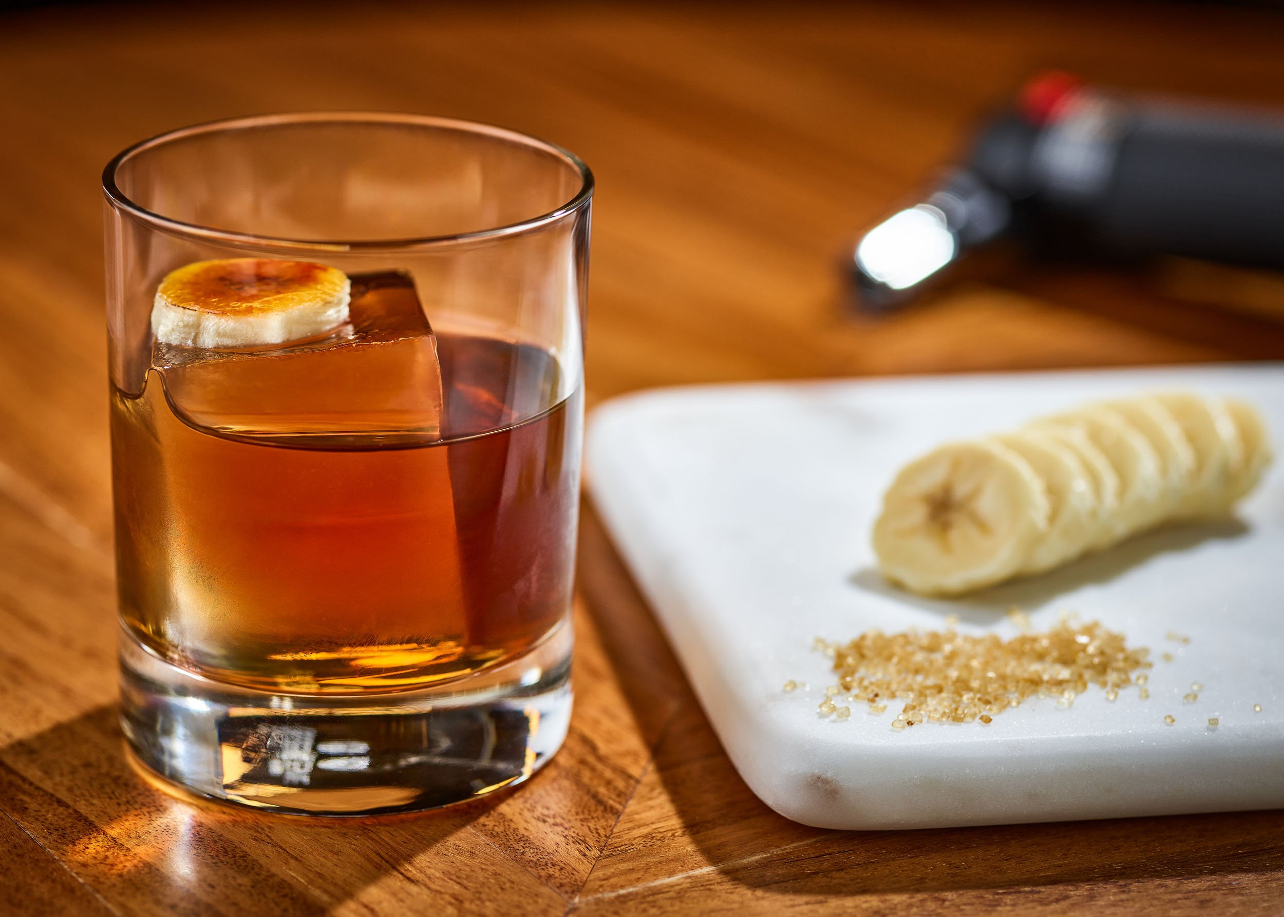 A glass of amber-colored liquor with a lemon slice on top, placed on a wooden surface. A white plate with sliced banana and crushed nuts or seeds is seen in the background.