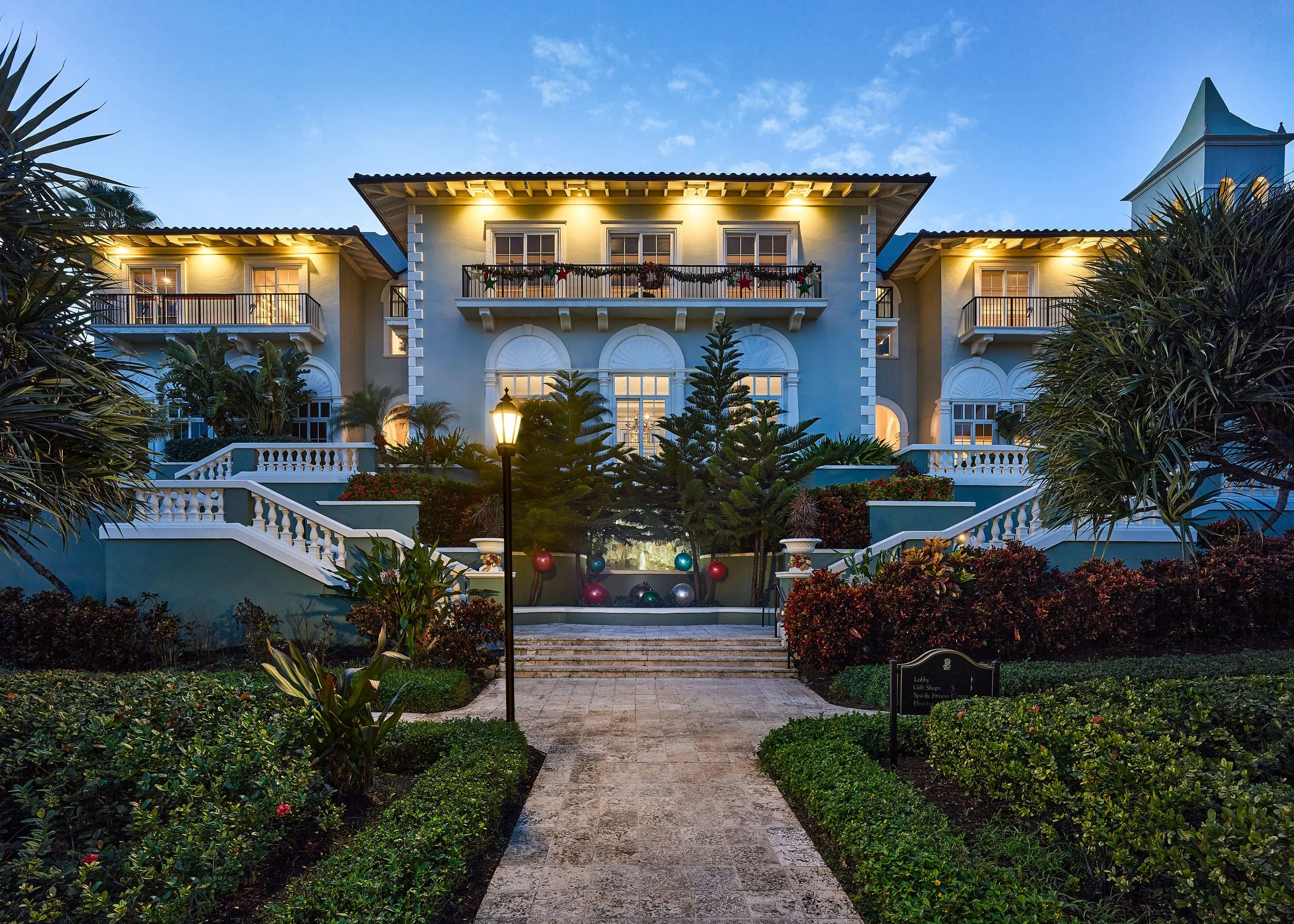 A large, elegant house with a well-kept garden in front, illuminated by exterior lights at dusk, featuring a central porch, two balconies, and Christmas decorations on the railing.