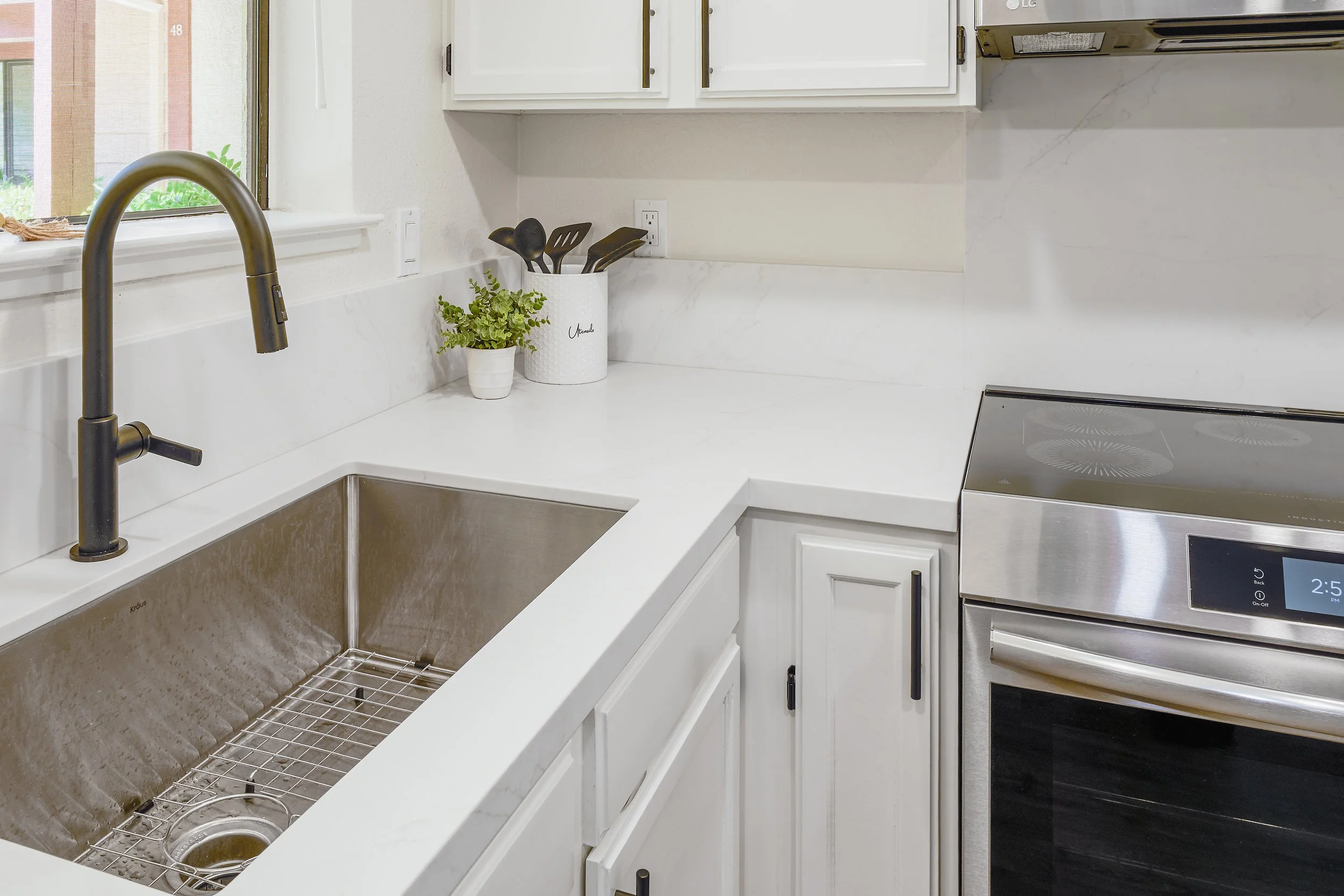A modern kitchen sink area with a black faucet, white cabinetry, a white countertop, a potted plant, and a utensil holder with black utensils. There is a window to the left and part of a stainless steel stove to the right.