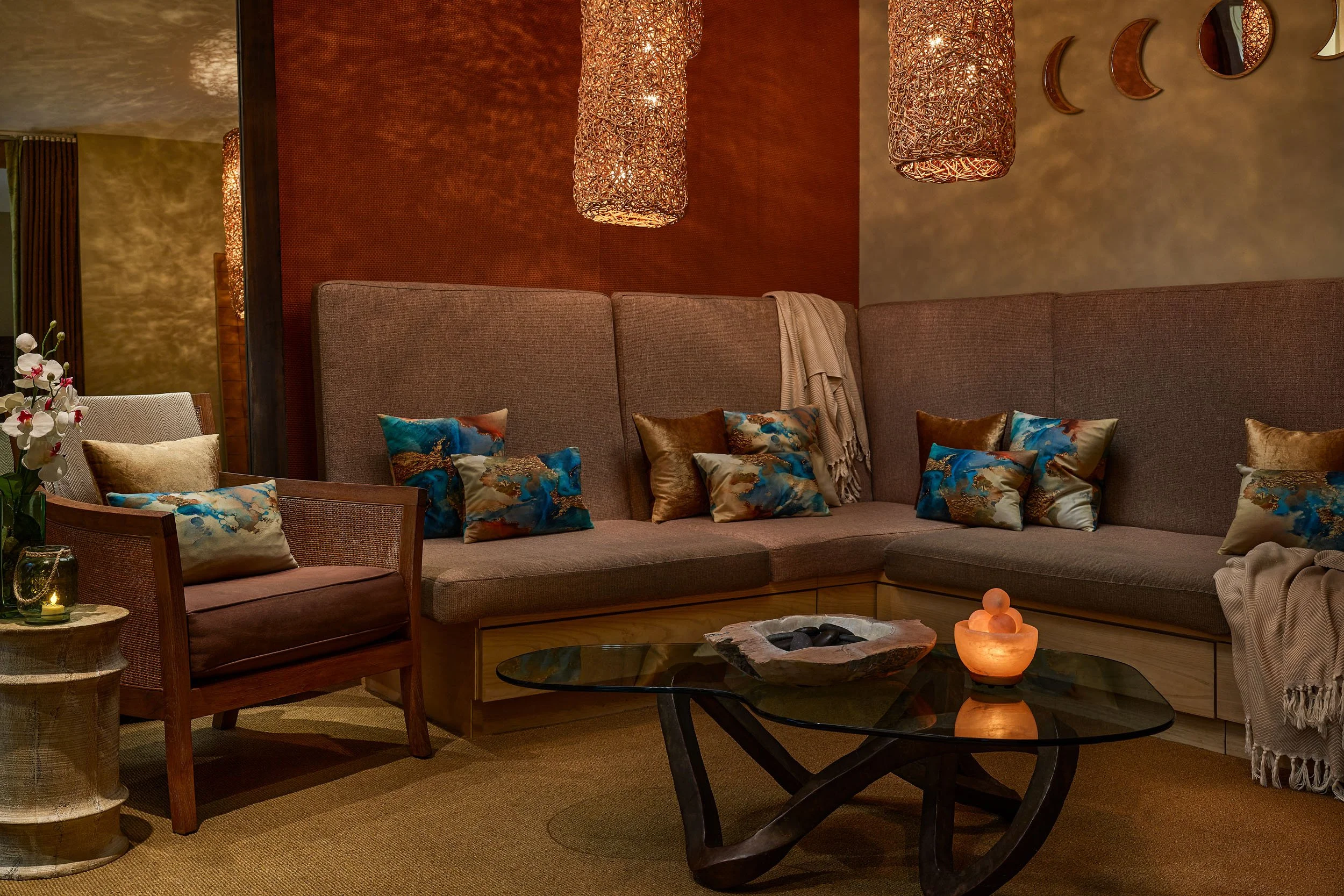 A cozy living room with a large sectional sofa adorned with colorful pillows, a round stone coffee table with a small decorative object, a wooden side table with a vase of orchids, and woven hanging lamps casting warm light and shadows.