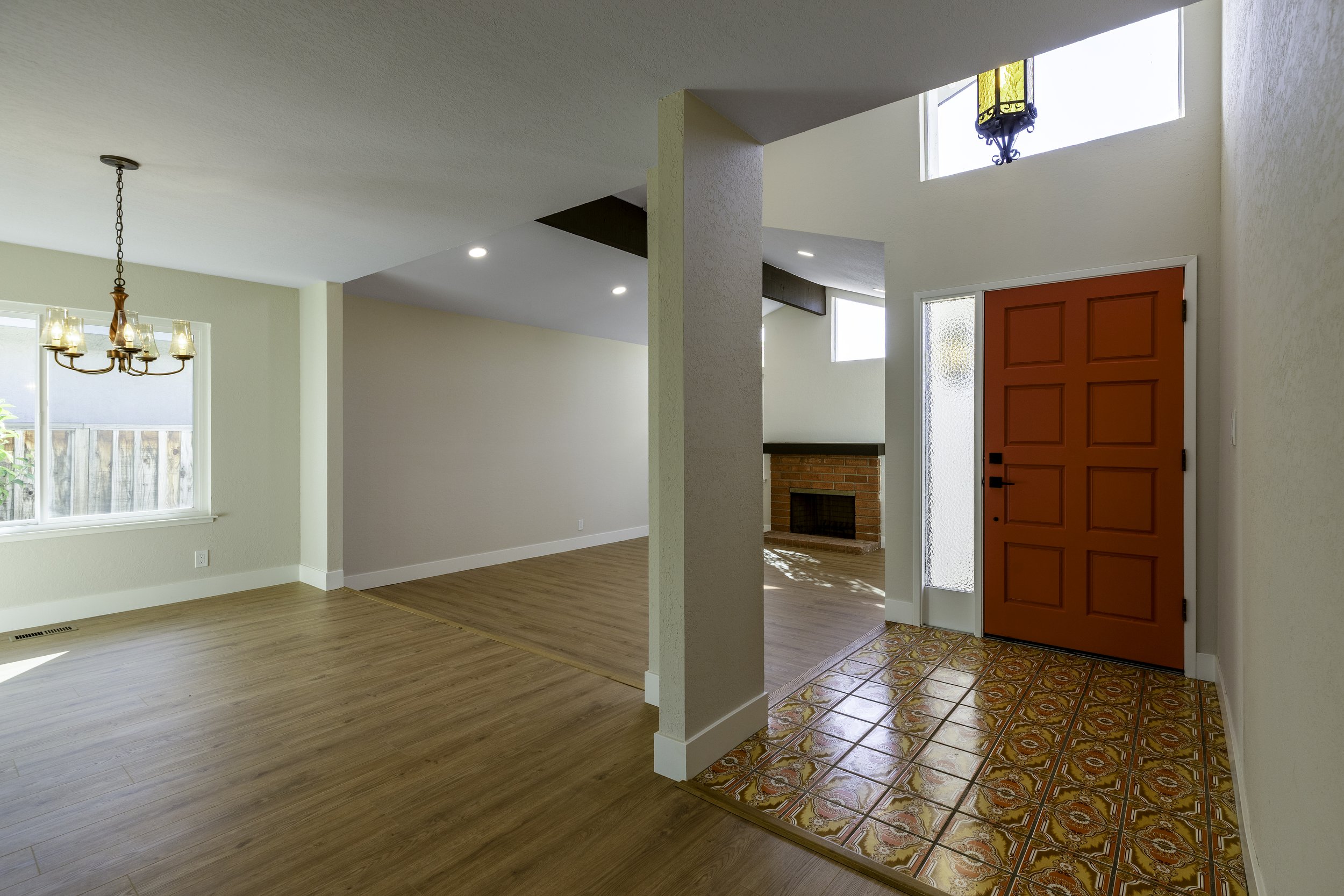 Interior view of a house with a red front door, wood and tile flooring, a brick fireplace, and sans-serif white walls, with windows letting in natural light.
