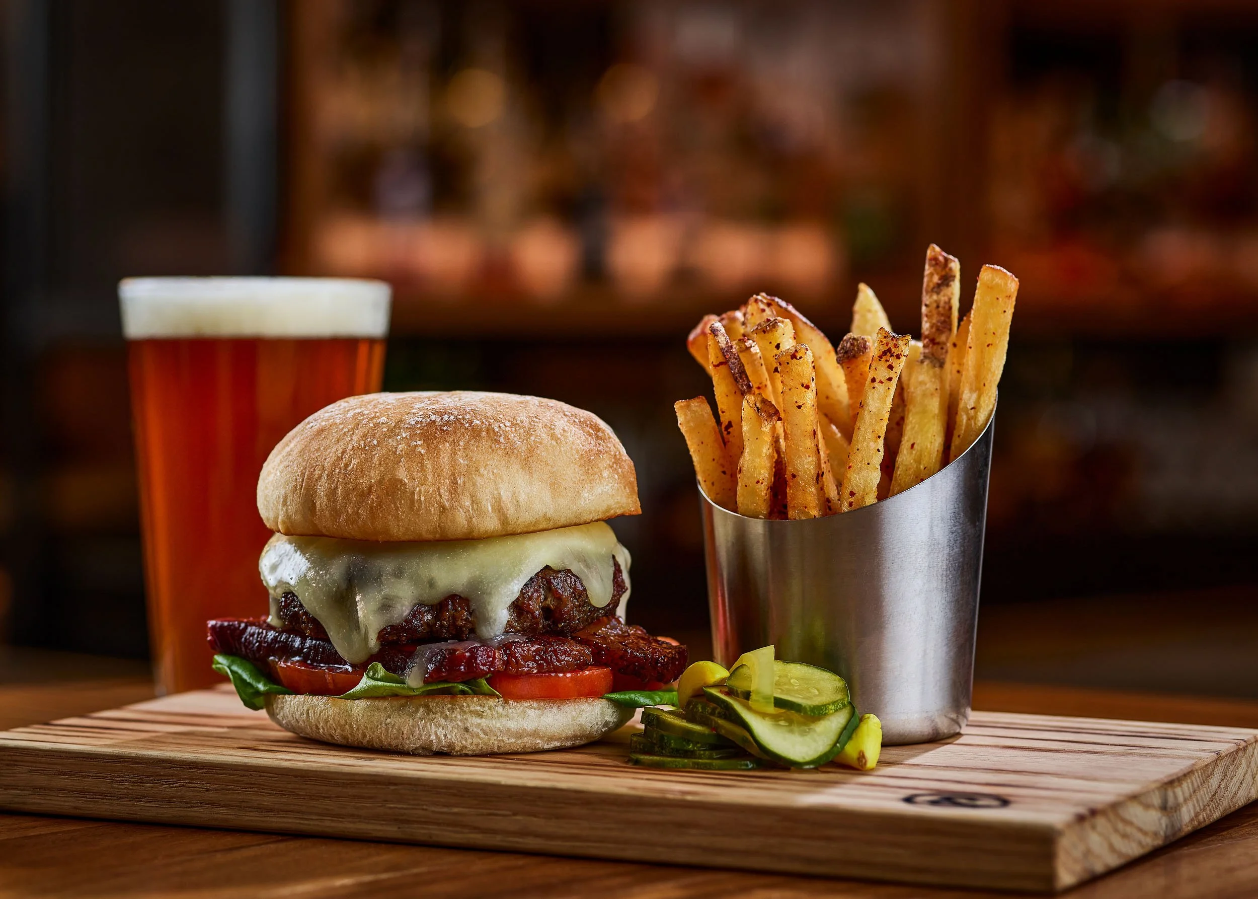 A burger with cheese, lettuce, tomato, and bacon inside a bun, accompanied by a side of sliced pickles, served on a wooden cutting board. In the background, there is a metal container of French fries and a glass of amber-colored beer with a foamy hea