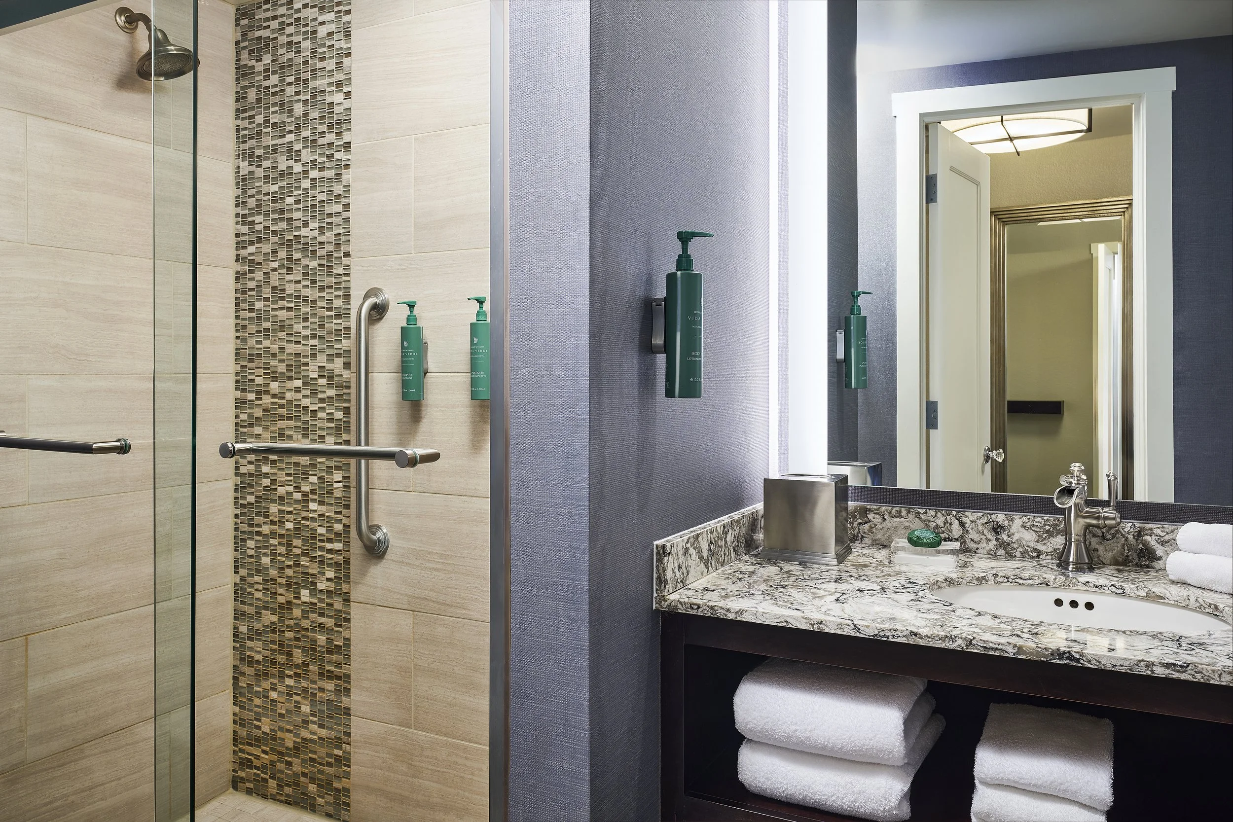 A hotel bathroom with a shower area featuring beige tiles and a decorative vertical strip of small mosaic tiles. The shower has metal grab bars, and there are three green bottles mounted on the wall. Next to the shower is a vanity area with a granite