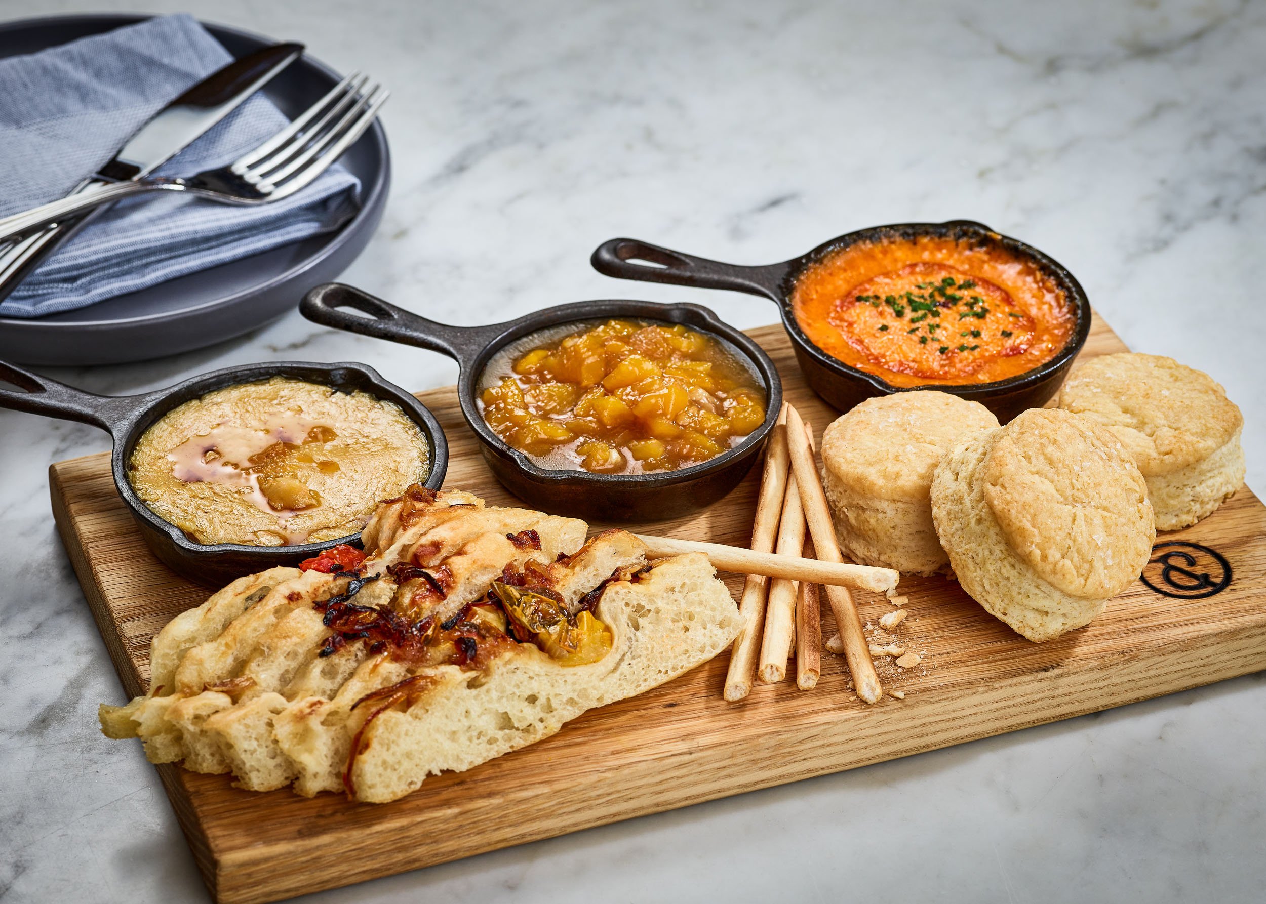 A wooden serving board with three small cast iron skillet dishes containing different types of cornbread and chili, along with breadsticks, on a marble table.