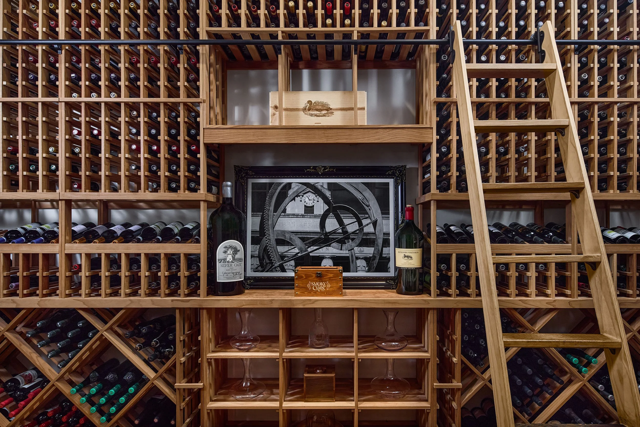 Wood wine rack filled with bottles, a framed clock and artwork, bottles of wine, glasses, and decorative items on the shelves.