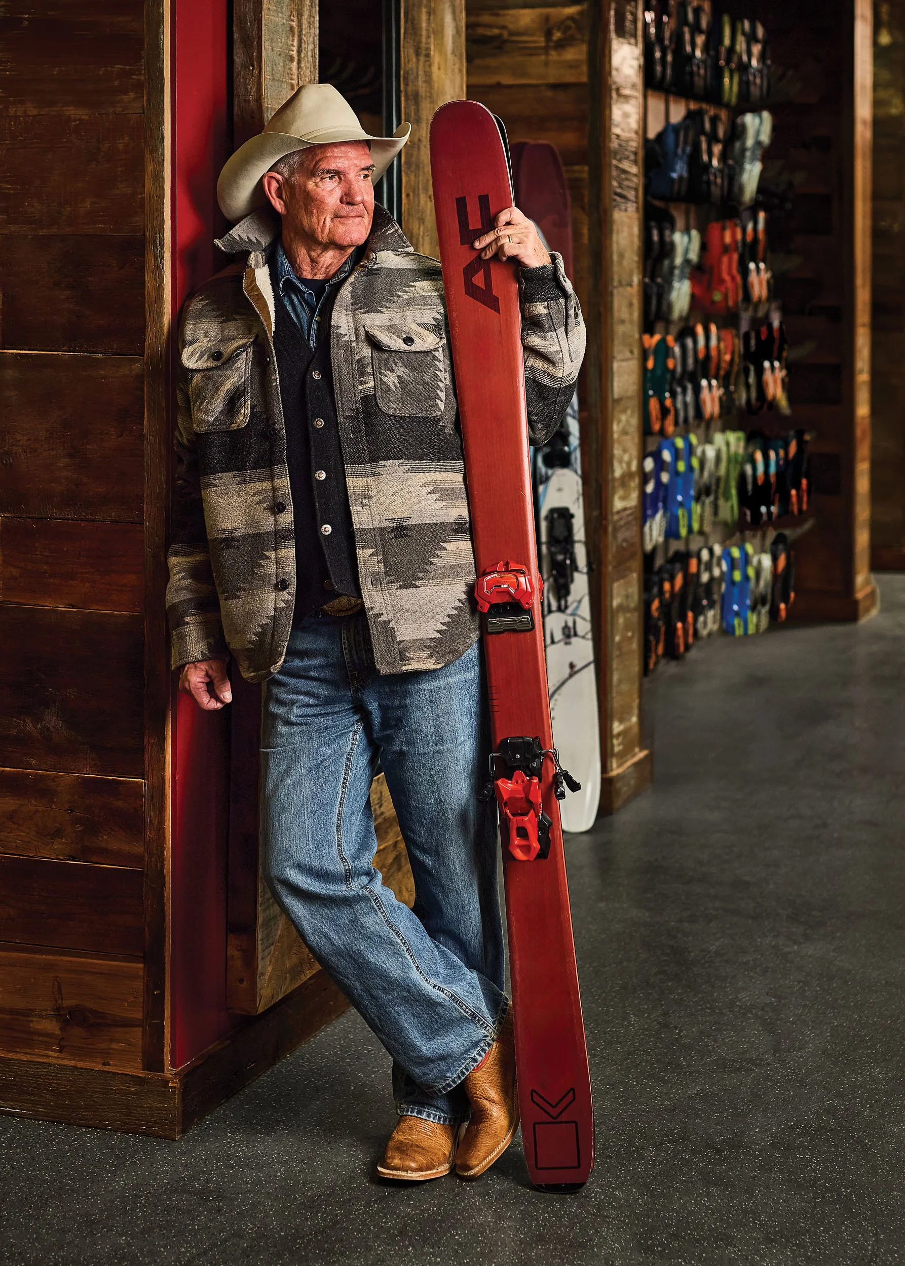 Older man wearing cowboy hat and cowboy boots, dressed in a patterned jacket and jeans, leaning against a wooden wall holding a red snow ski with the brand name 'Atomic' and a snowboard in the background, inside a store with skiing gear on shelves.