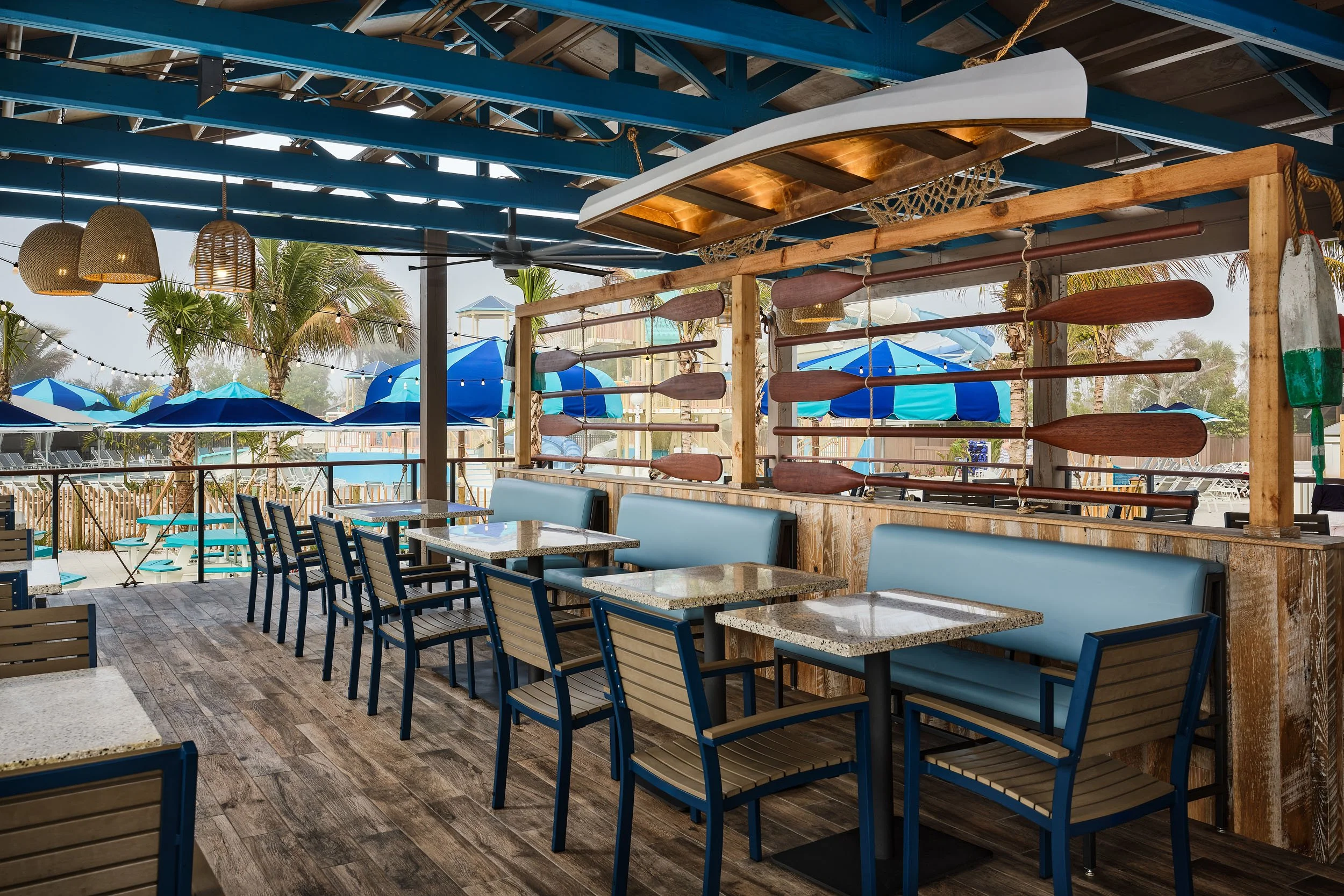 Outdoor seating area of a restaurant or café with tables, blue chairs, hanging wicker lamps, palm trees, and blue umbrellas visible outside. There is a wooden surfboard-shaped decoration with paddles on a wooden frame.