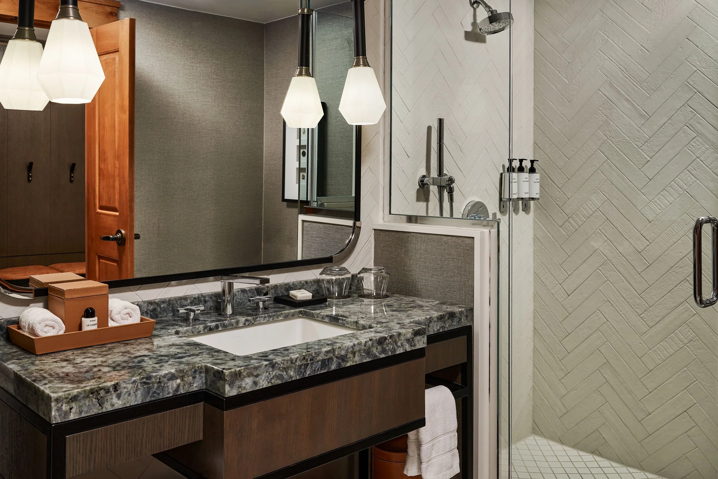 A modern bathroom featuring a granite countertop with a sink, a large mirror, wall-mounted soap dispensers, and a glass-enclosed shower with a rain showerhead. There are three pendant lights hanging above the sink area. The decor includes textured wa