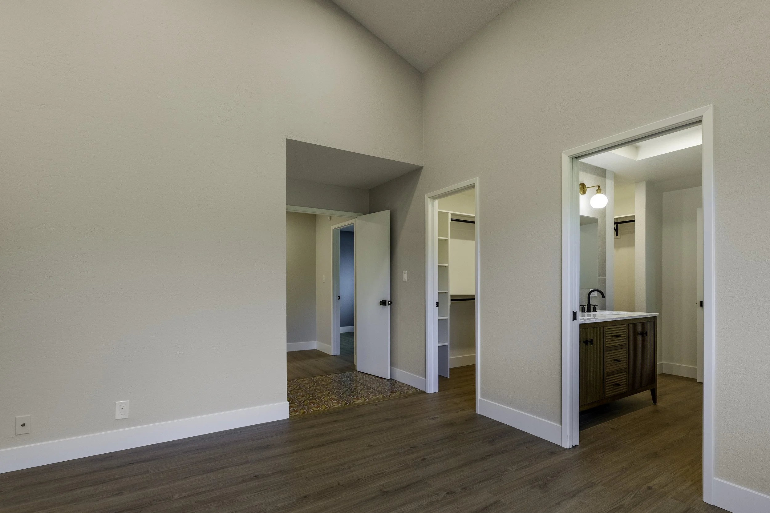 Empty interior room with white walls, wood floor, and open doorway to a small bathroom with a wooden vanity, black faucet, and white walls.