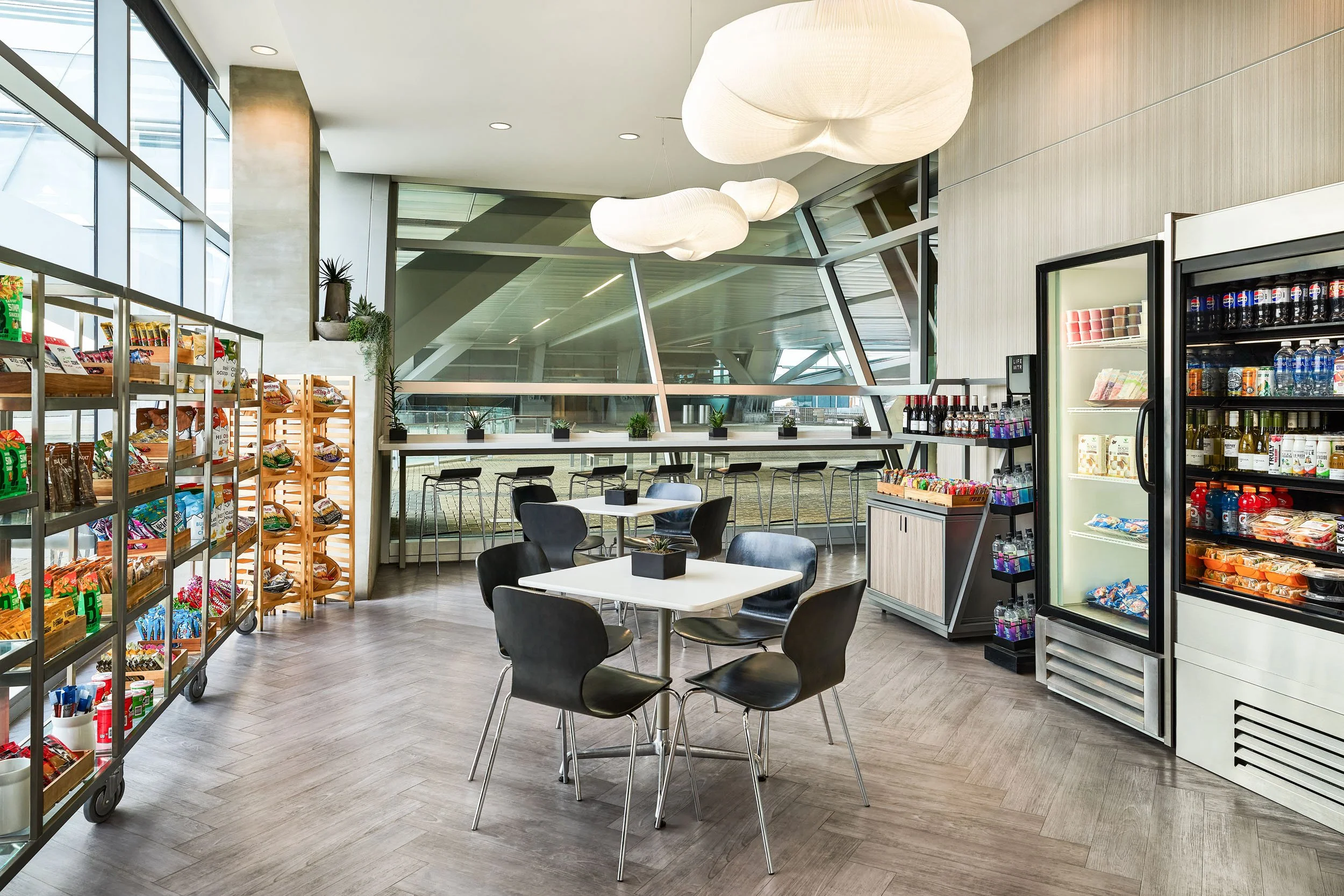 Modern indoor dining area with tables, chairs, snack shelves, and a refrigerated beverage display, illuminated by large ceiling pendant lights.