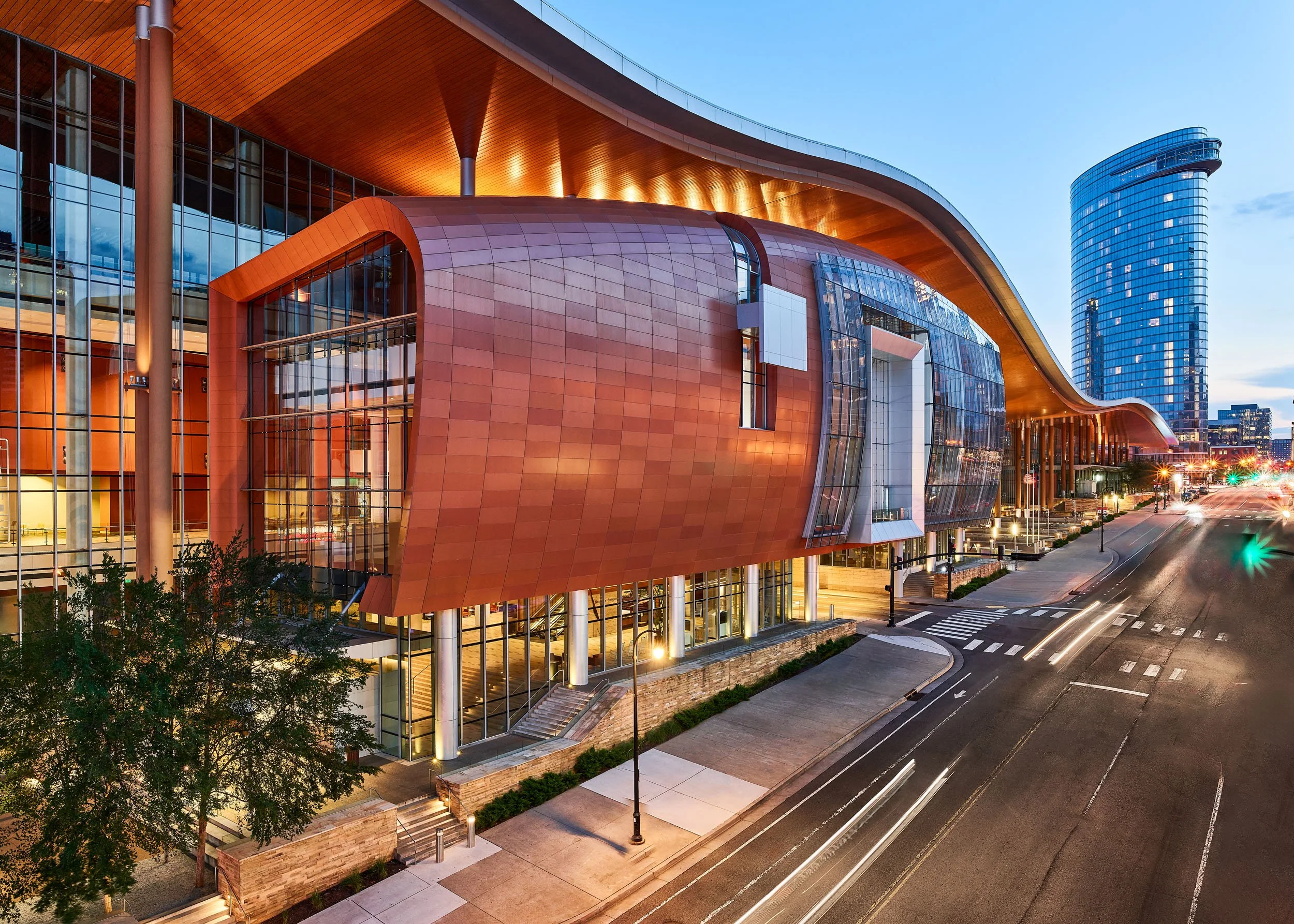 Nighttime exterior view of a modern building with a curved copper-colored roof, glass walls, and a tall blue skyscraper in the background. Street lights and traffic with blurred car lights are visible.