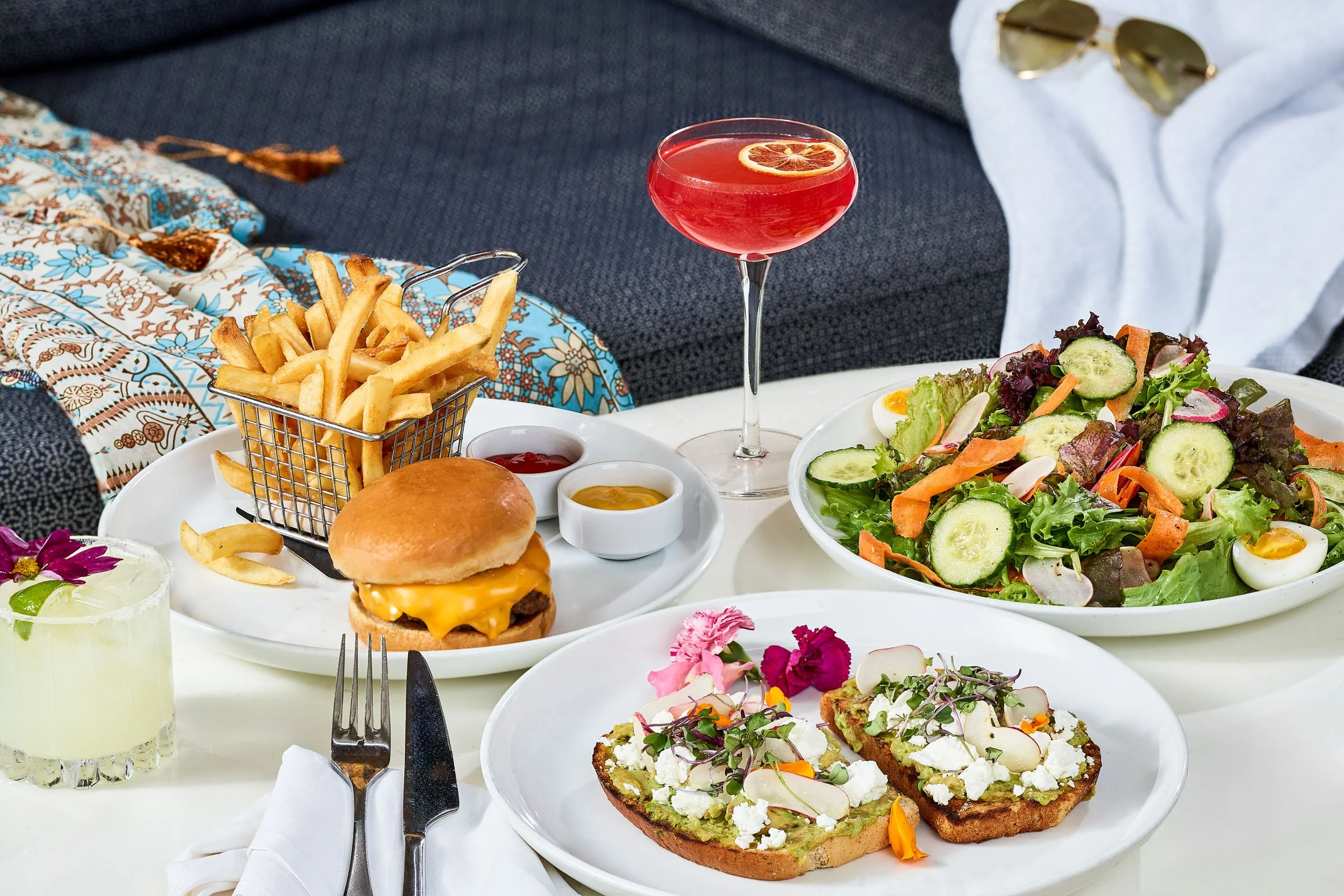 A table filled with various dishes including a cheeseburger, French fries, a salad, toasted bread with cheese and vegetables, a glass of a red drink with a lemon slice, and small bowls of condiments. There are also sunglasses and a white napkin on th