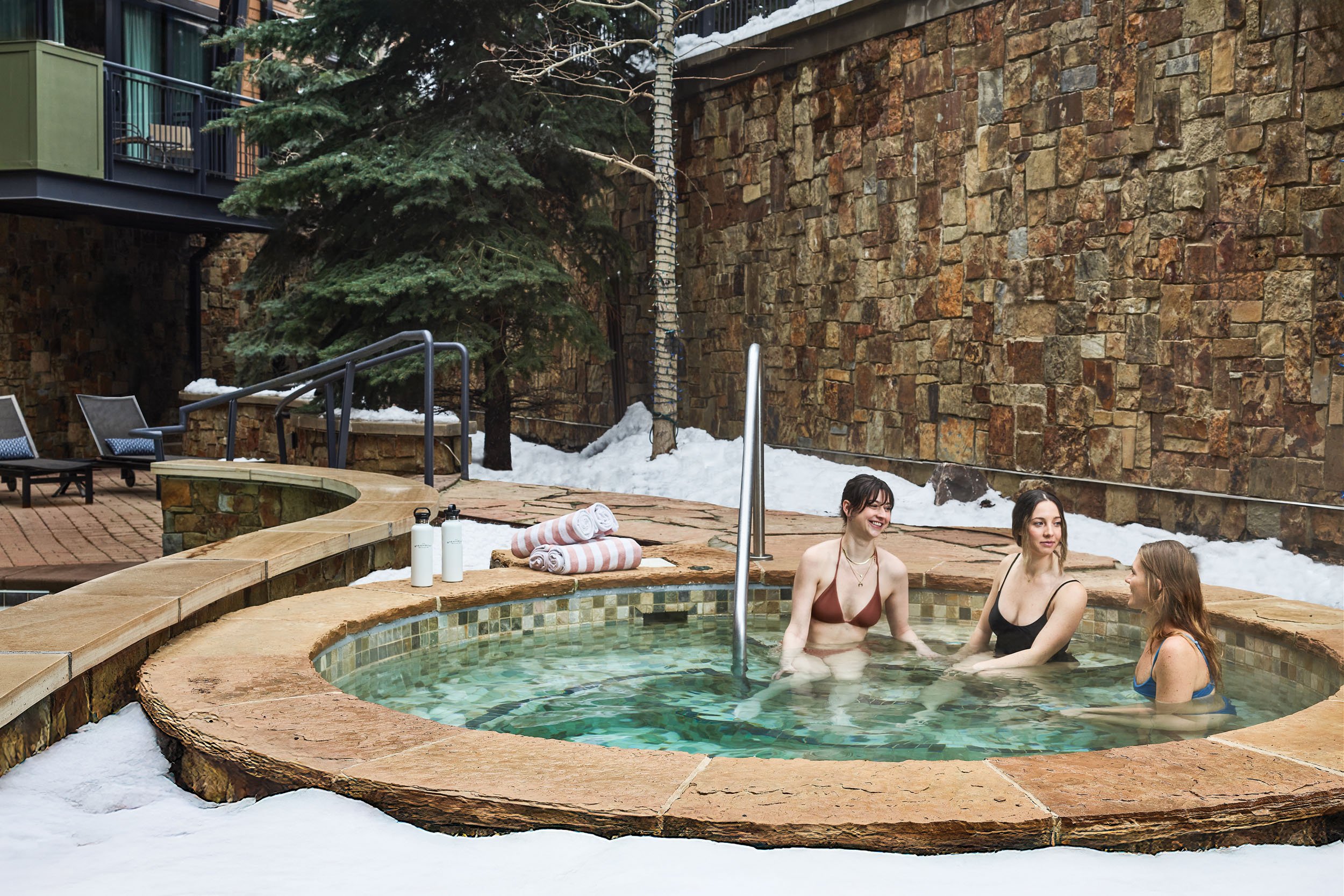 People sitting in a hot tub outdoors with a stone wall, snow, and trees in the background.