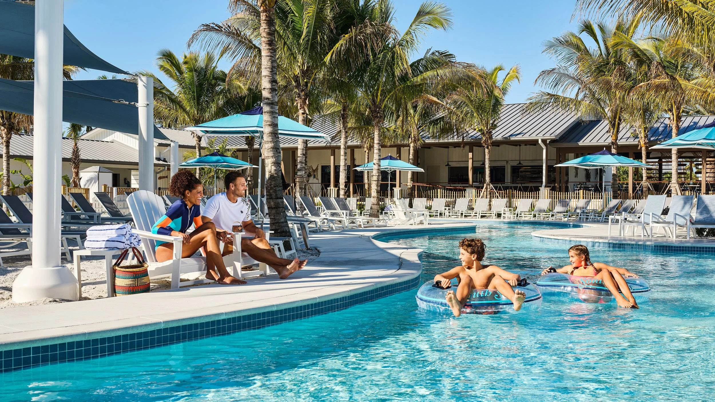 People relaxing and playing in a swimming pool at a tropical resort with palm trees, lounge chairs, umbrellas, and a building in the background.
