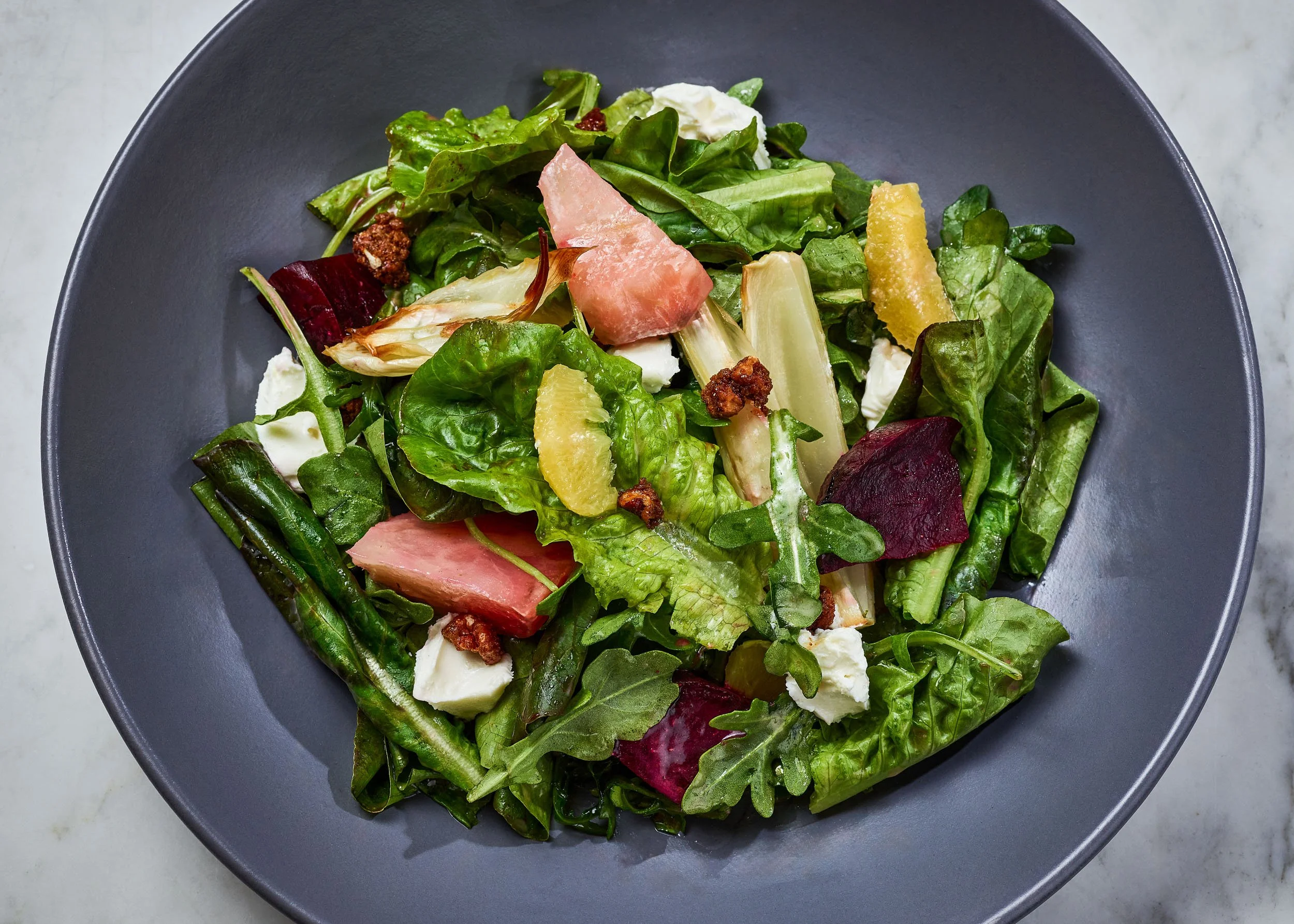 Fresh mixed greens salad with slices of watermelon, crumbled cheese, and balsamic glaze on a black plate.