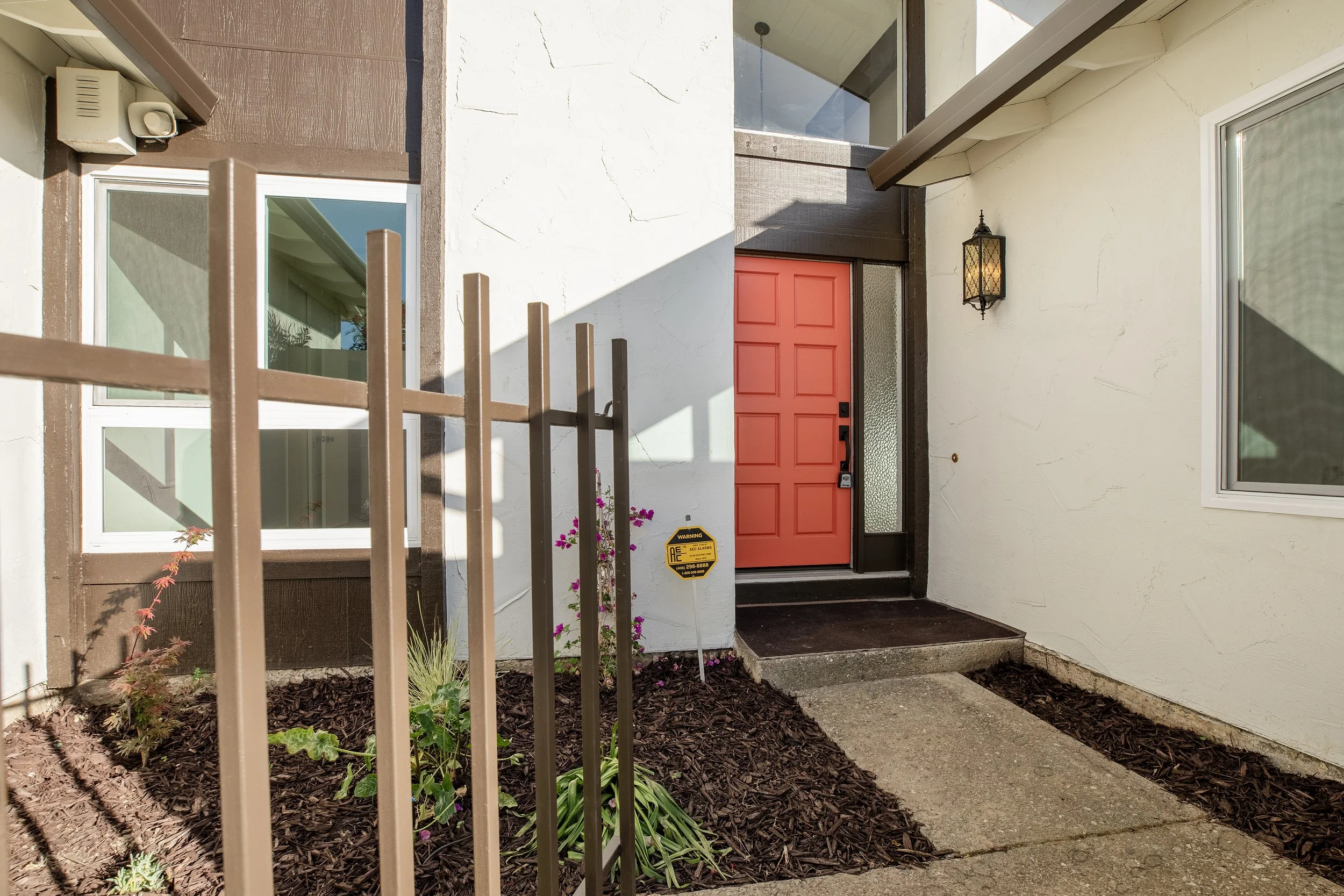 Front entrance of a house with a bright orange door, decorative lantern, small garden bed with plants, and window on beige wall.