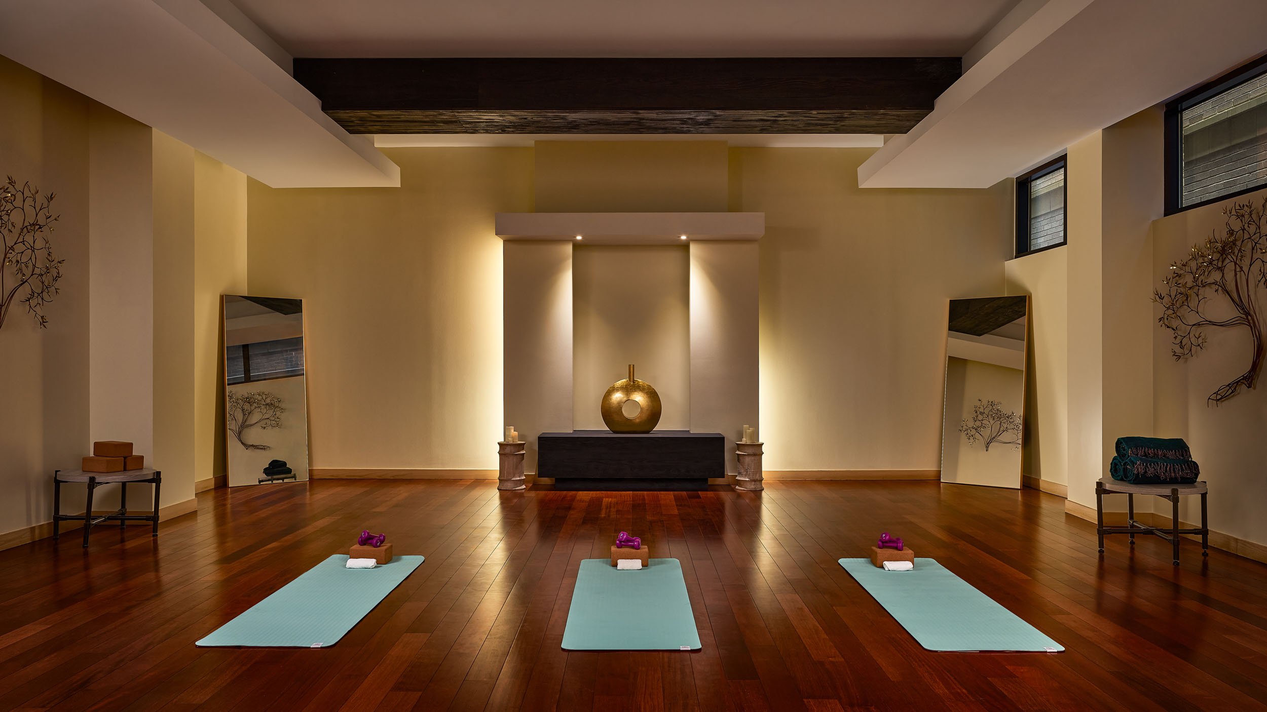A yoga or workout room with four light blue yoga mats, each with a purple dumbbell, a small towel, and a wooden block on top. The room features wood flooring, two mirrors leaning against the walls, decor including trees on the wall, and a central alt