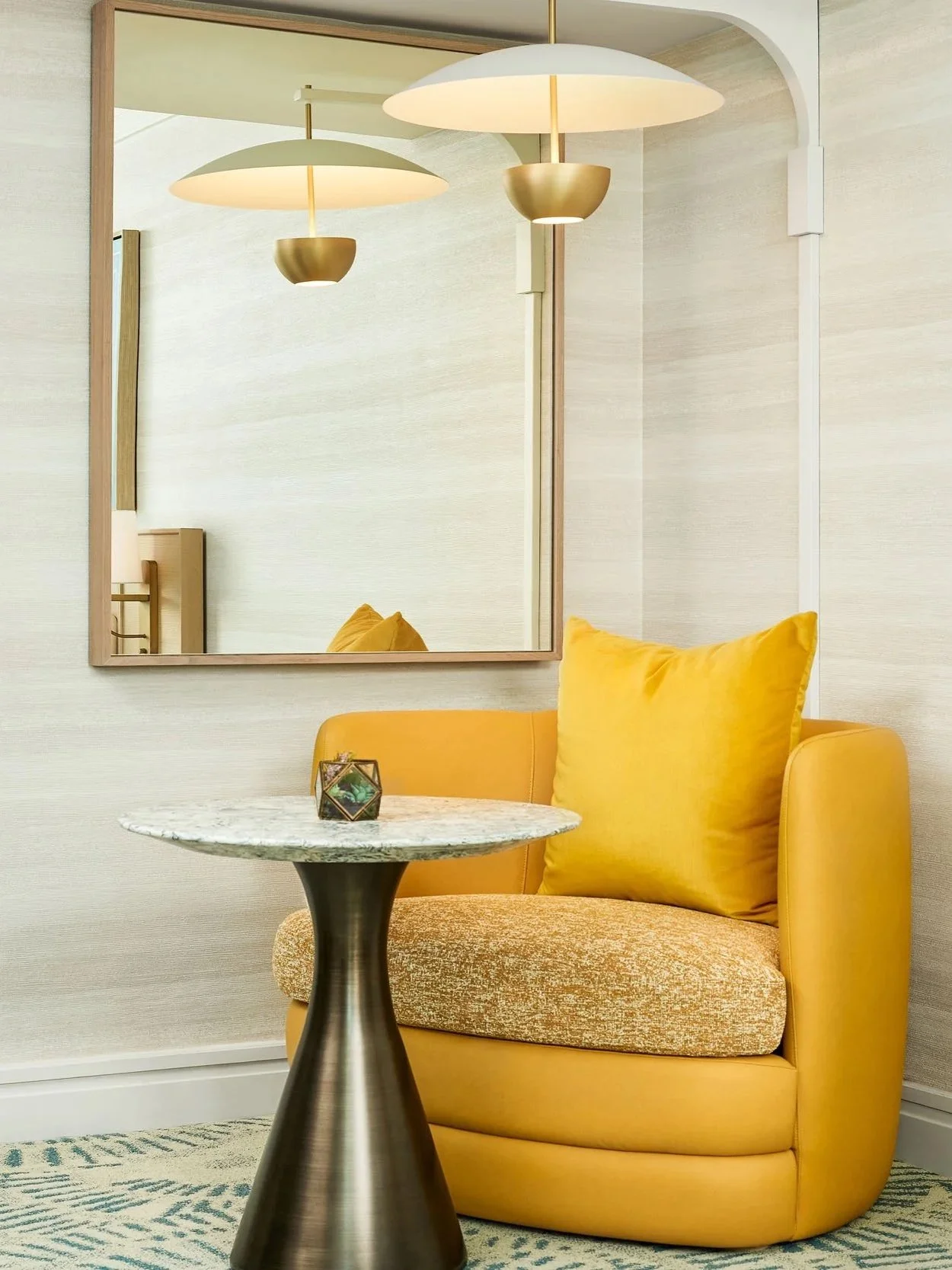 A yellow armchair with a matching yellow pillow, a small round marble-topped side table, a decorative glass terrarium, a large mirror on the wall, and modern pendant lights reflected in the mirror in a cozy, well-lit room.