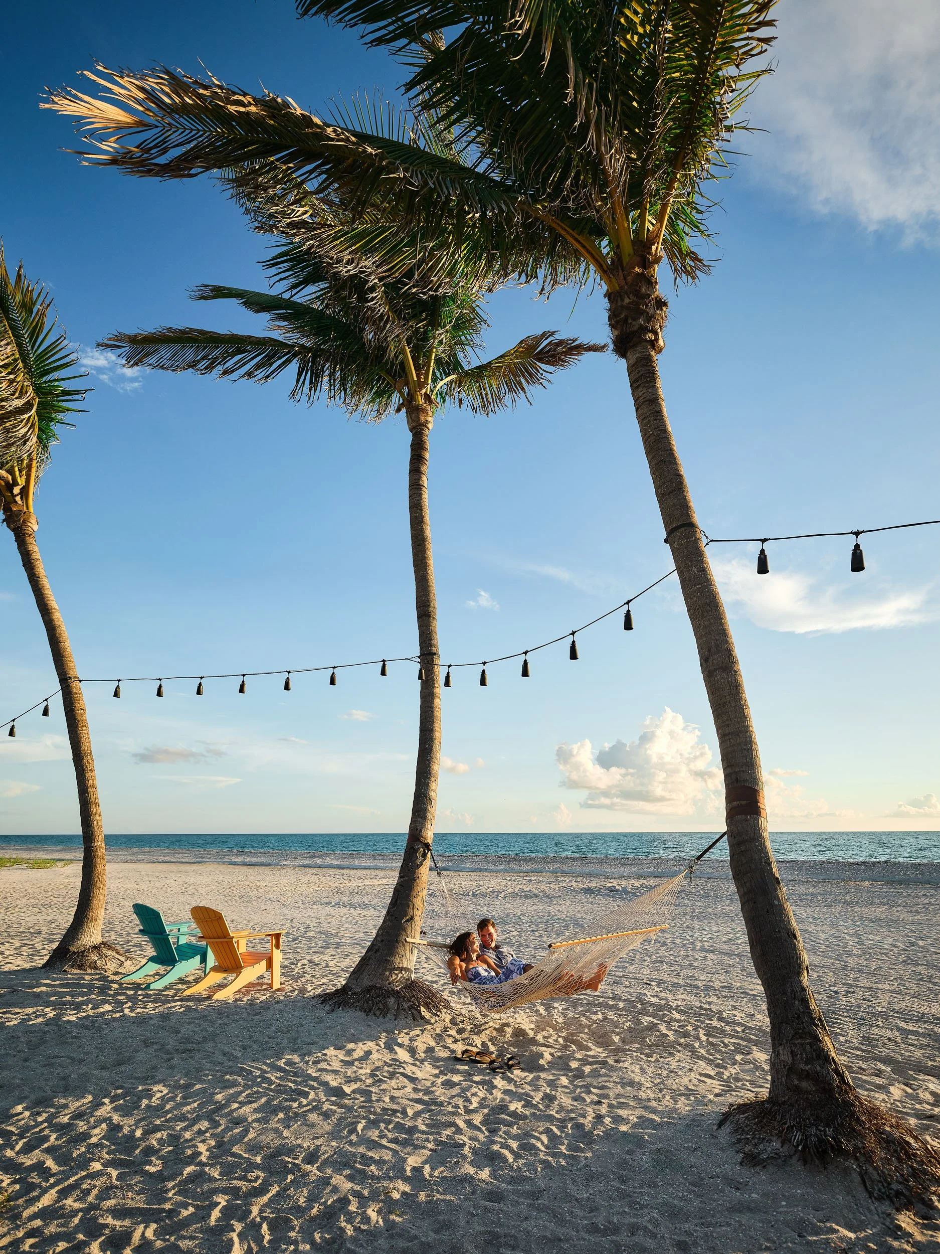 A couple relaxing in a hammock between two palm trees on a beach with Adirondack chairs nearby, ocean in the background, clear sky with some clouds, and string lights hanging between the palm trees.